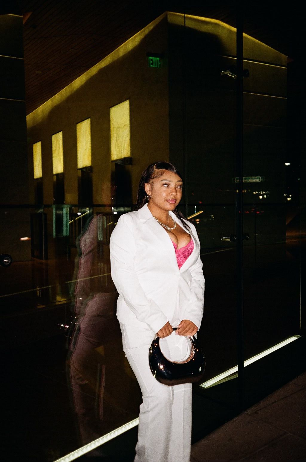 Woman in white suit with pink top holding a black handbag, standing in a modern interior with dark walls and lighting.