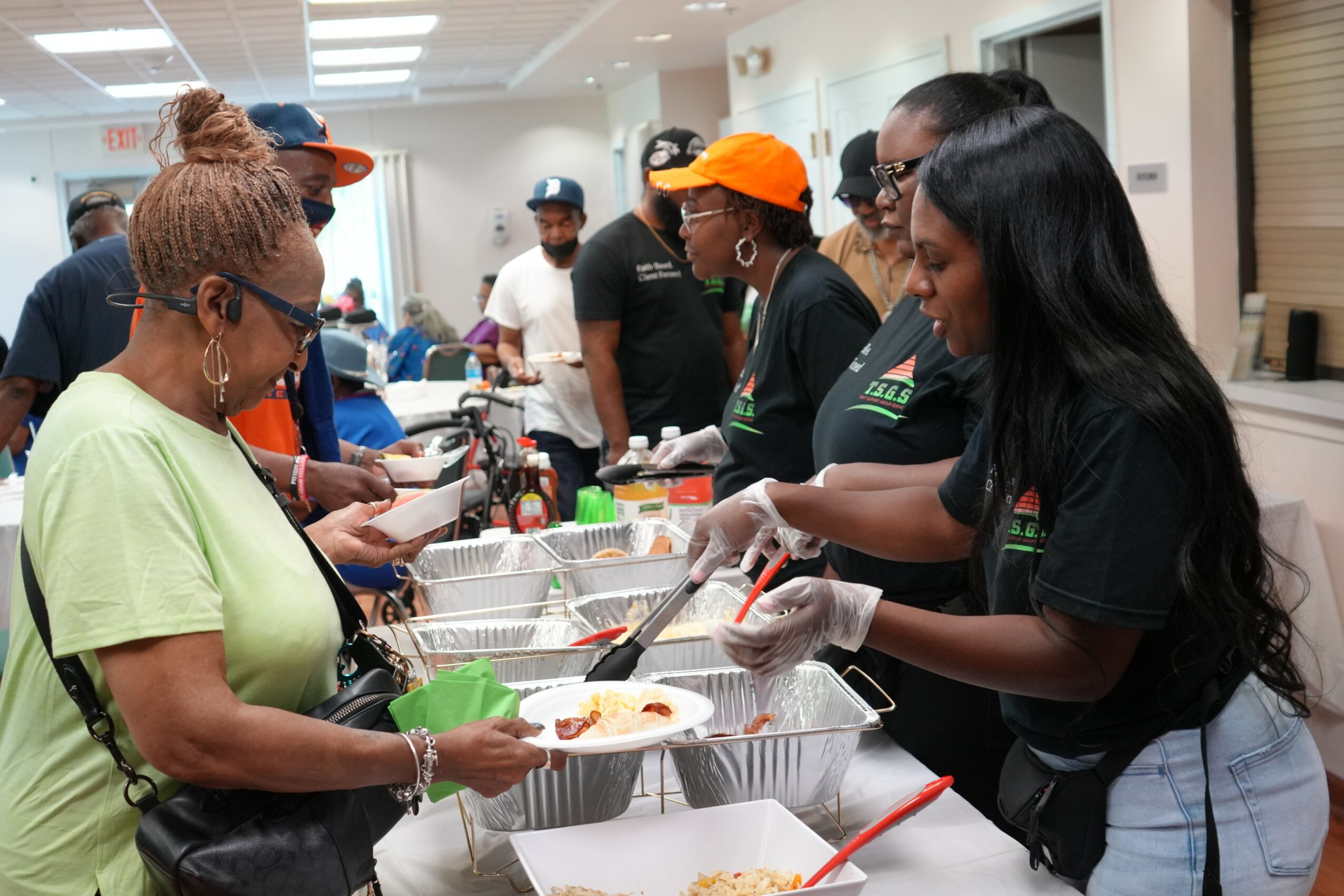 People in a line at a food service counter, serving food to a woman in a green shirt, with others waiting behind.