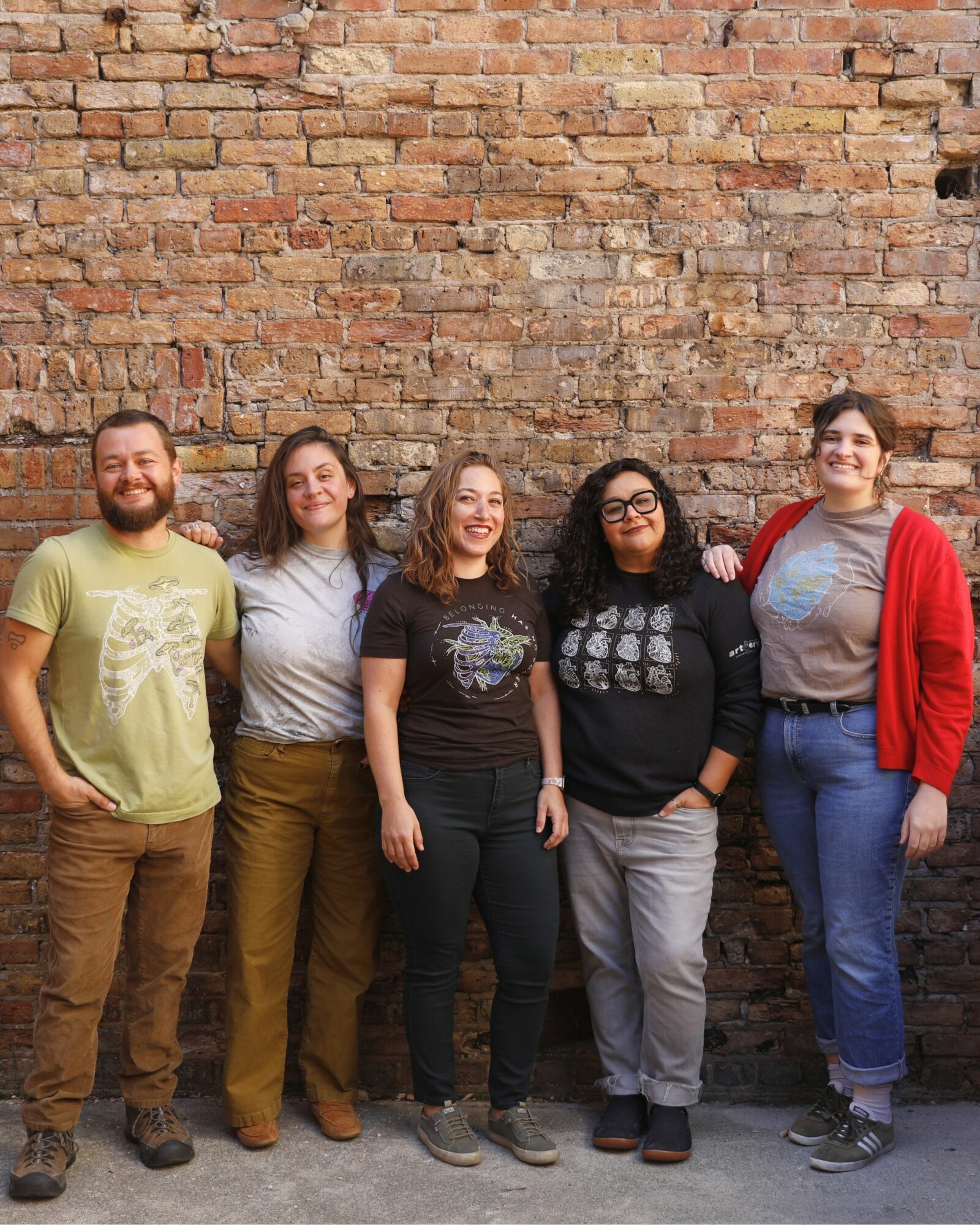 Five people standing in front of a brick wall, smiling, with arms around each other.