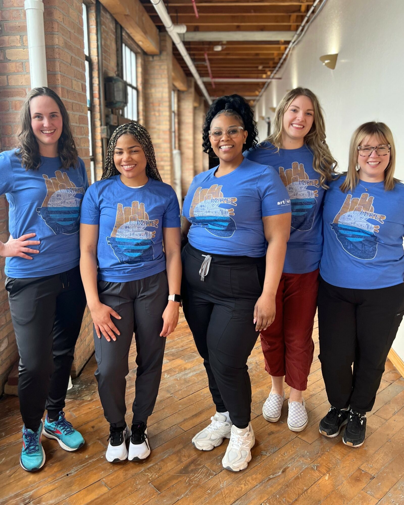 Five women standing in a hallway, smiling, wearing matching blue T-shirts with a graphic design, and casual pants.