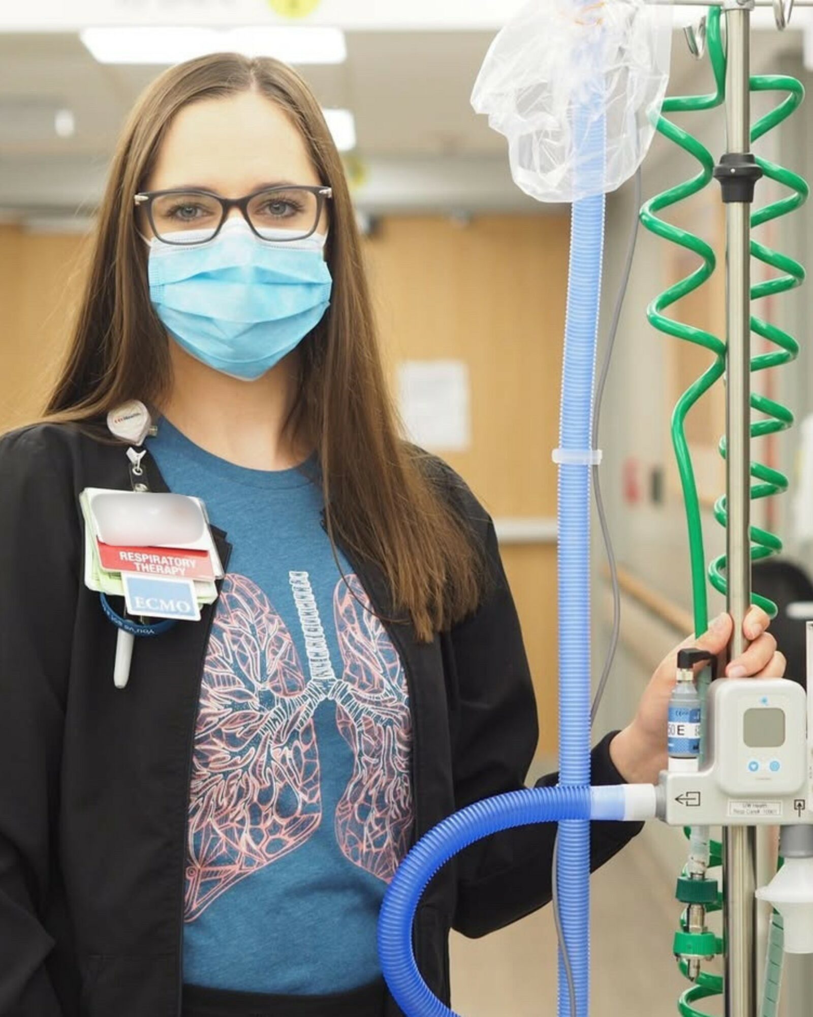 Young woman wearing glasses and a face mask in a medical setting, holding medical equipment, with a hospital background.