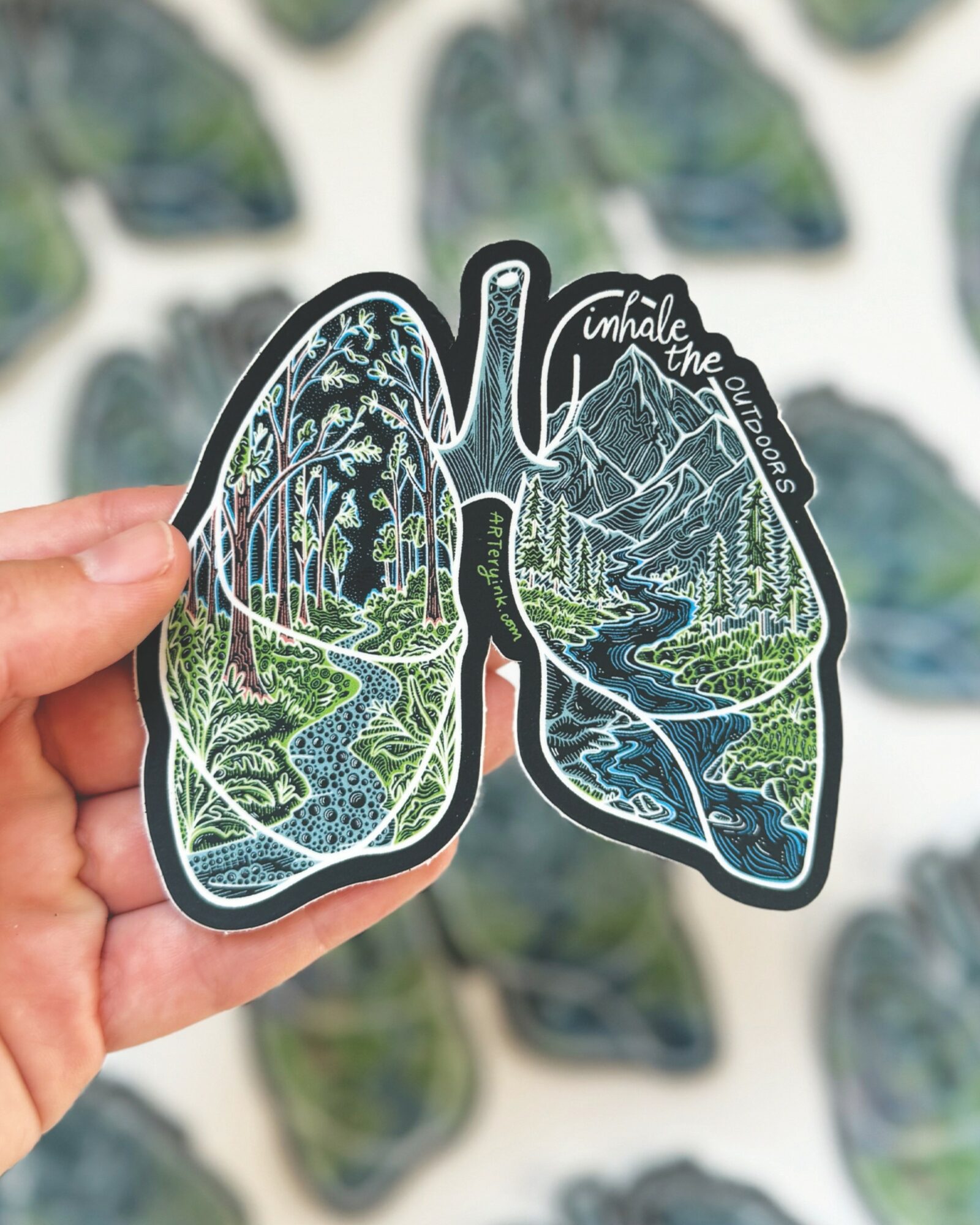 Sticker of lungs with landscape of mountains, trees, river, and sky inside, held by a hand.