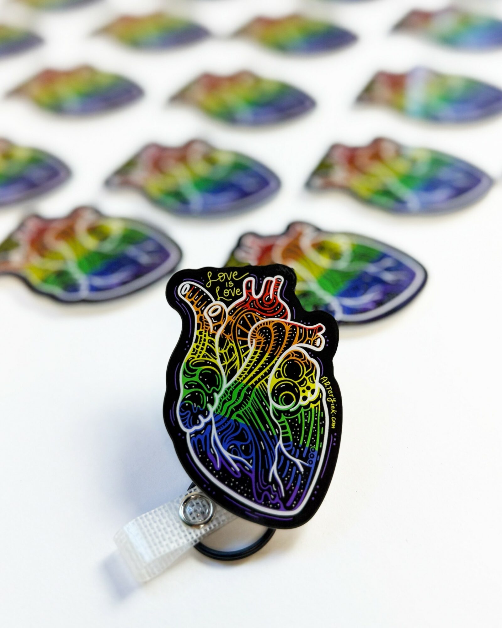 Colorful rainbow heart-shaped sticker with a detailed anatomical heart illustration, placed on a white surface with many similar stickers in the background.