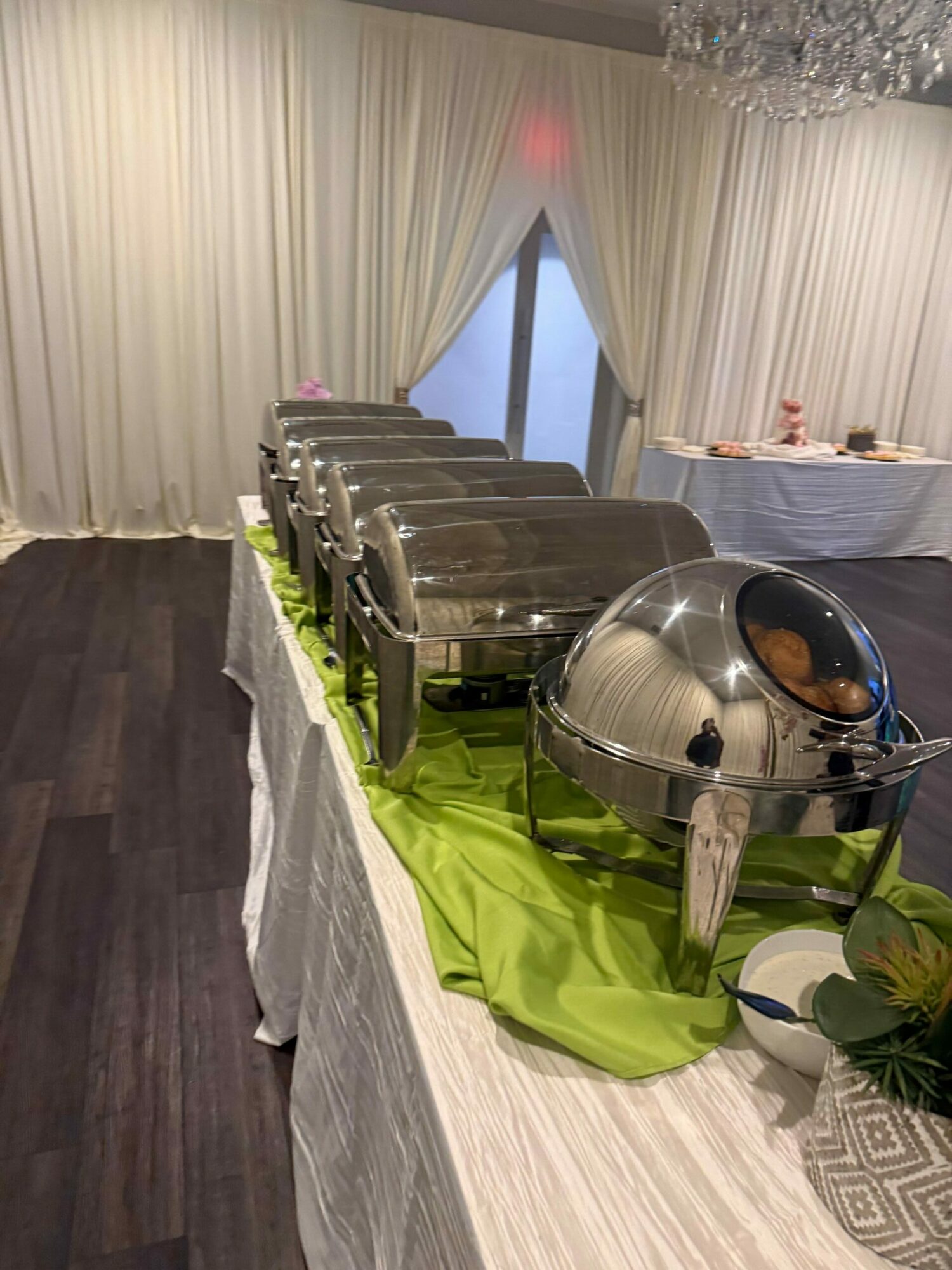 Buffet table with chafing dishes, green cloth, and decorative items, viewed from the side, with curtains and window in background.