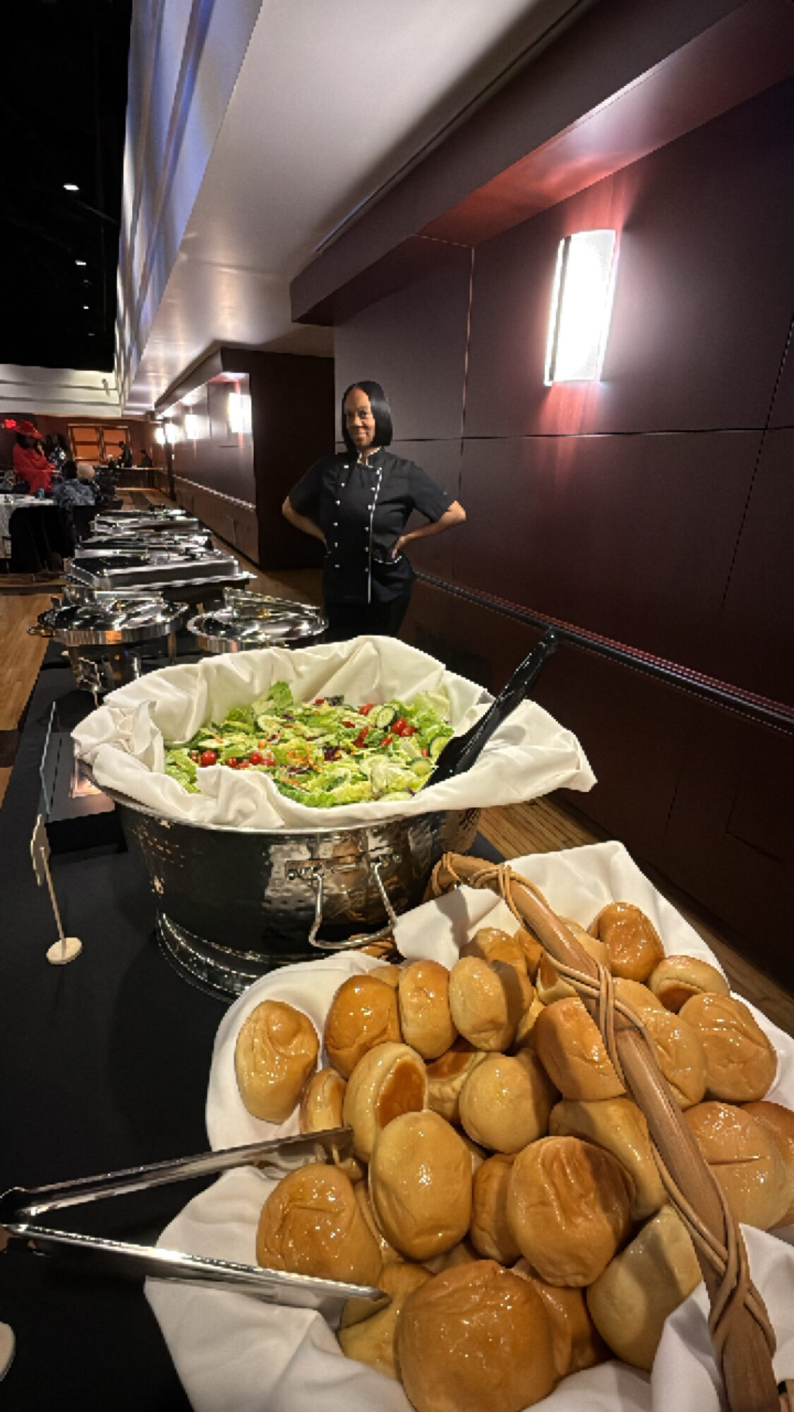 Person standing behind a buffet table with a large bowl of salad and a pile of bread rolls, in a dimly lit room.