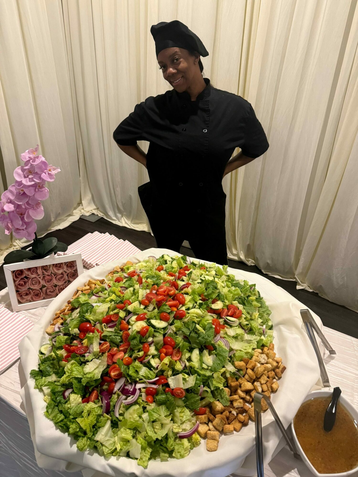 Person standing next to a large salad on a table, with a bowl of dressing and a drink nearby.