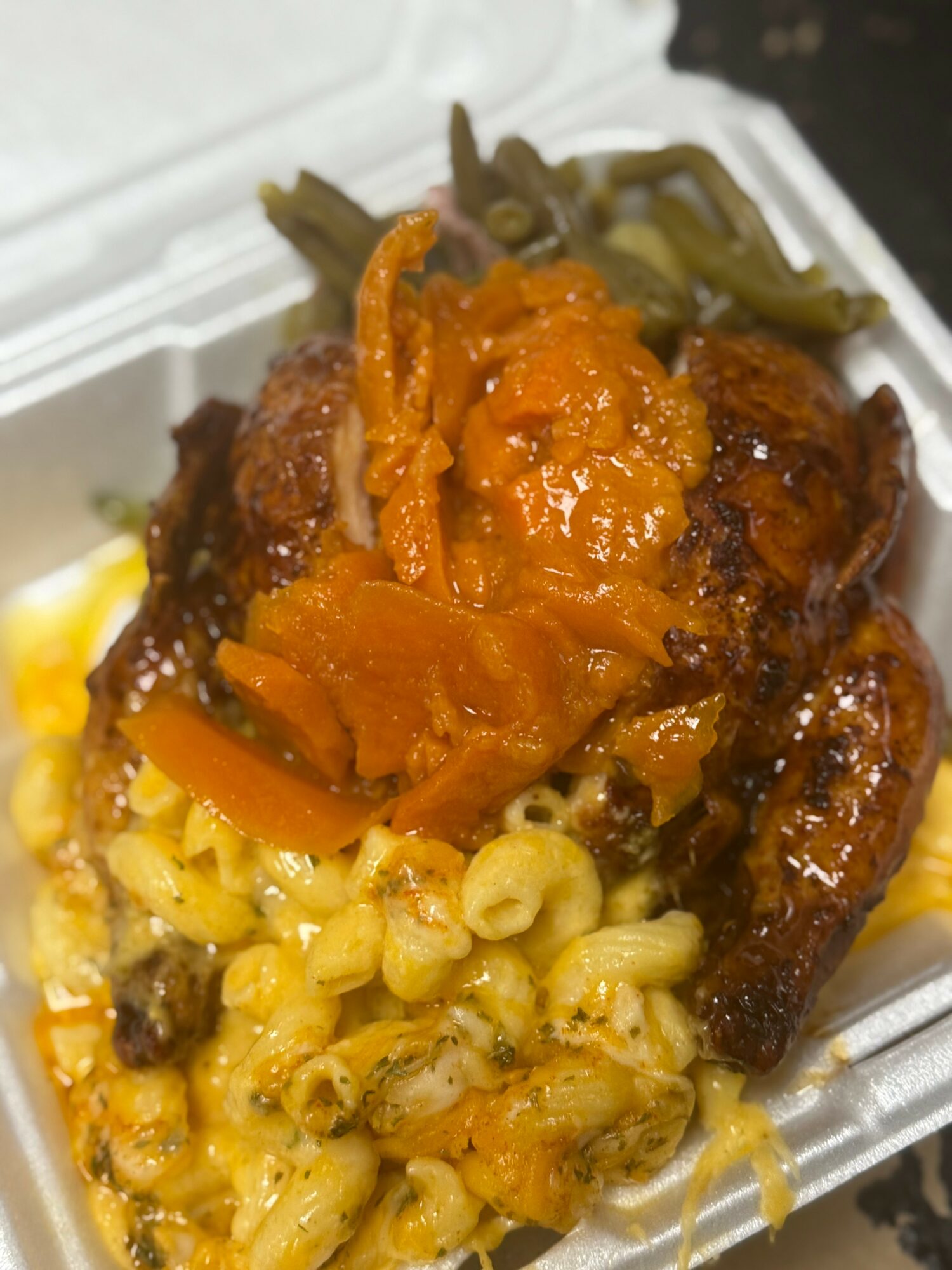 Fried chicken with macaroni and cheese, topped with orange sauce and pickles in a foam container.