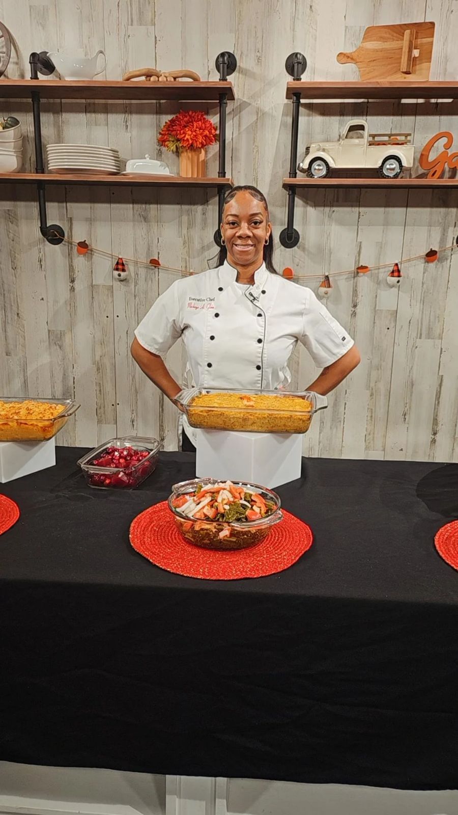 Chef standing behind a table with food, smiling, in a kitchen setting with shelves and decorations