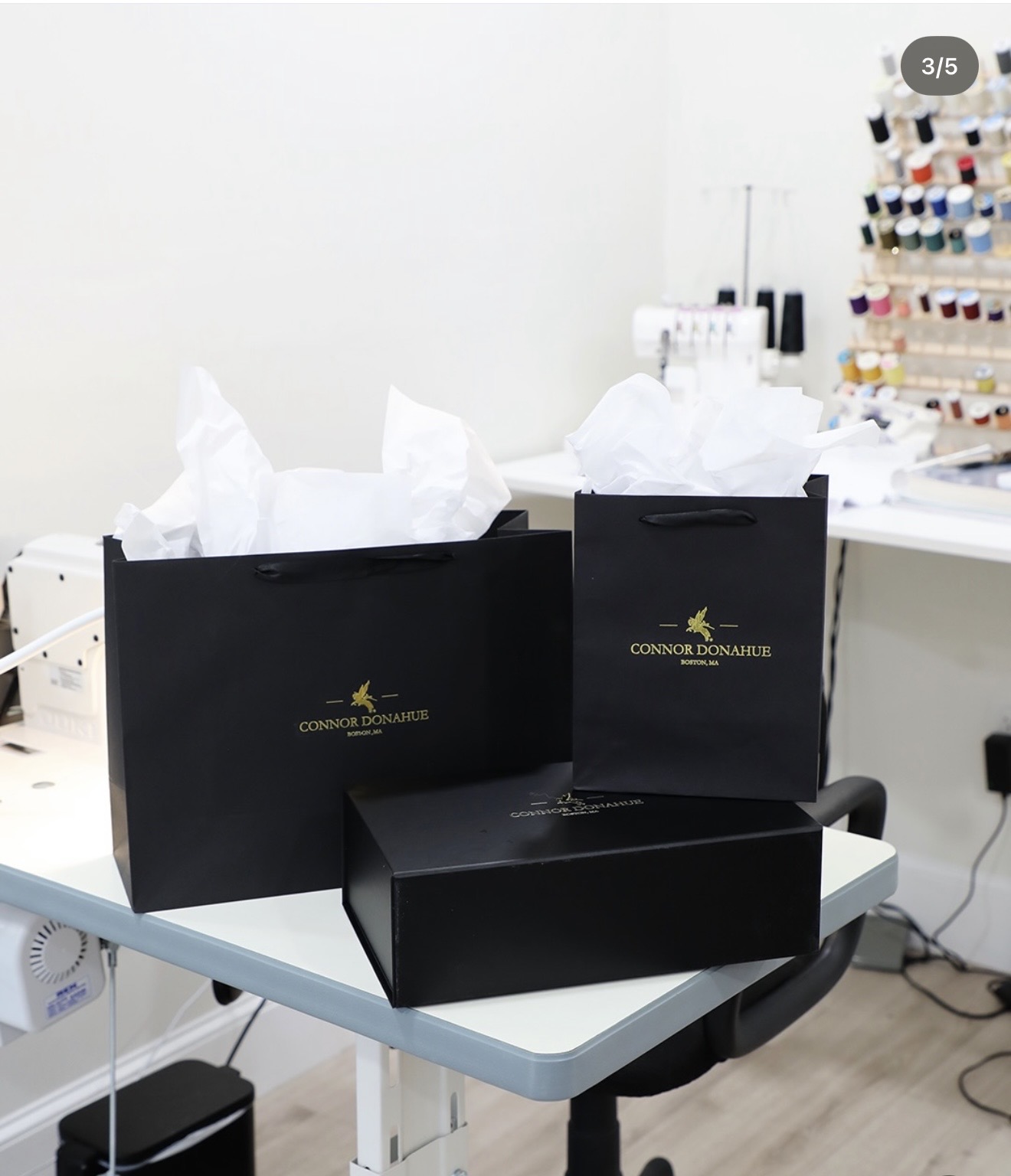 Two black shopping bags with tissue paper and a black box on a table, sewing supplies in background