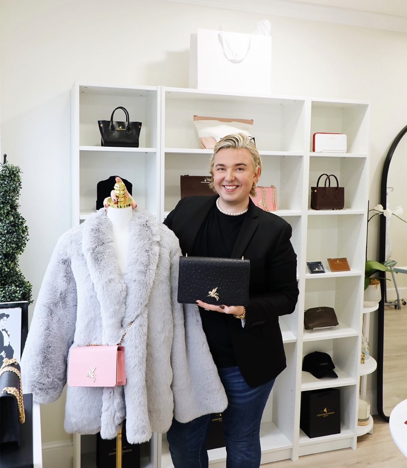 Person with short blonde hair smiling, holding a black clutch, standing in front of white shelves with bags and accessories.