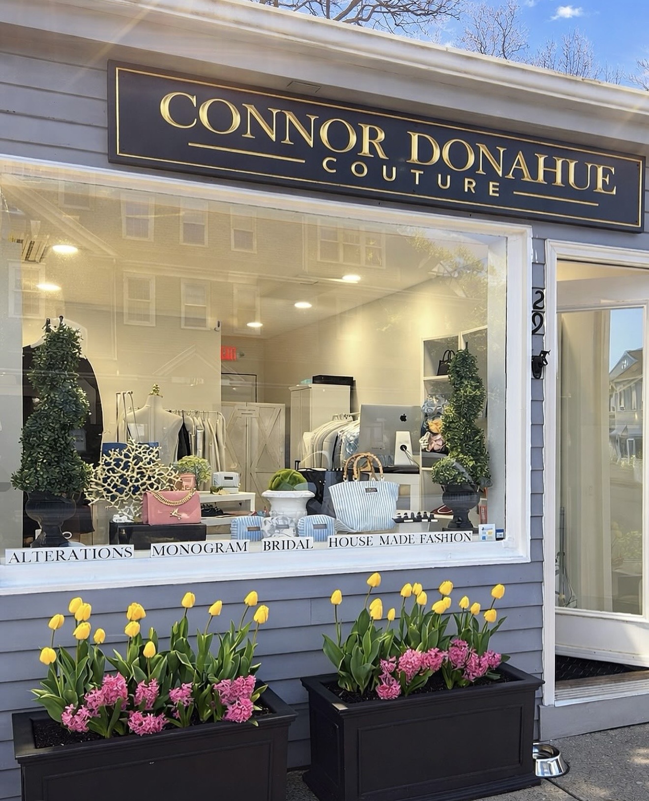 Storefront with Connor Donahue Couture sign, window display with accessories, and flower planters with yellow tulips and pink flowers outside.