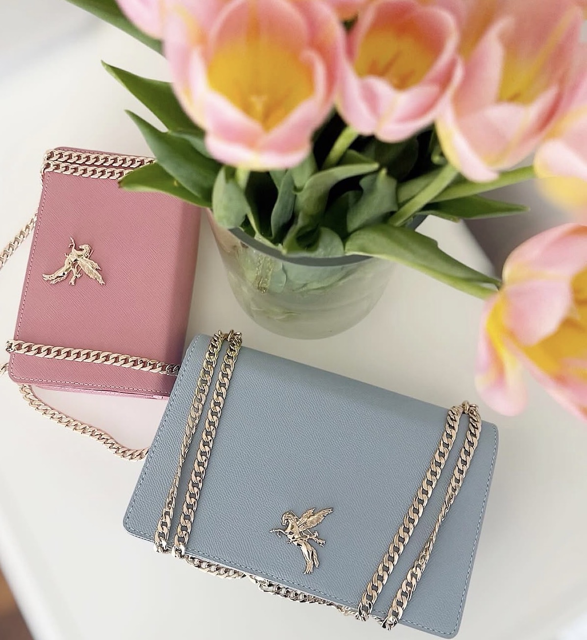 Pink and blue handbags with chain straps, a bouquet of pink tulips in a glass vase, on a white surface.