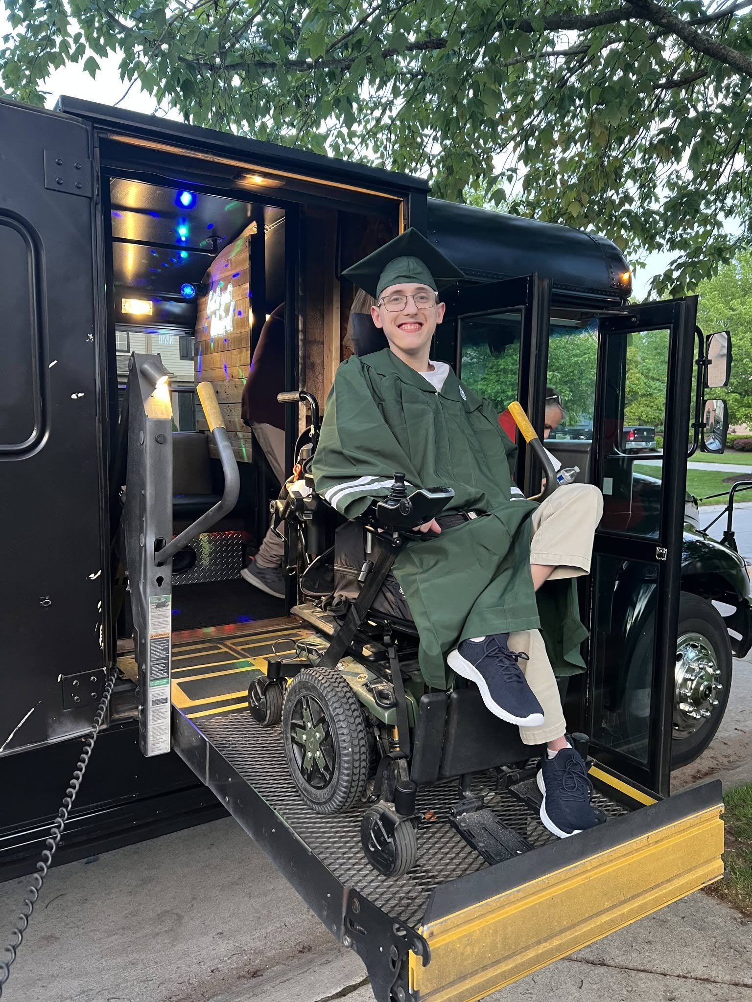 Person in a green graduation gown and cap sitting in a wheelchair on a bus with open door, outdoors with trees in background.