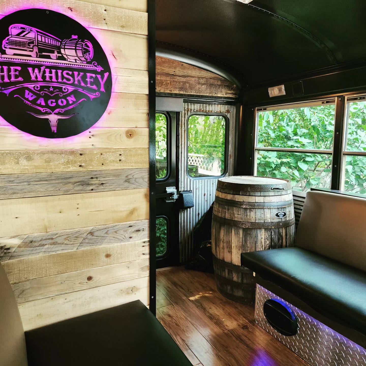 Interior of a bus with wooden walls, a barrel table, and seating, with a window view of green trees outside.