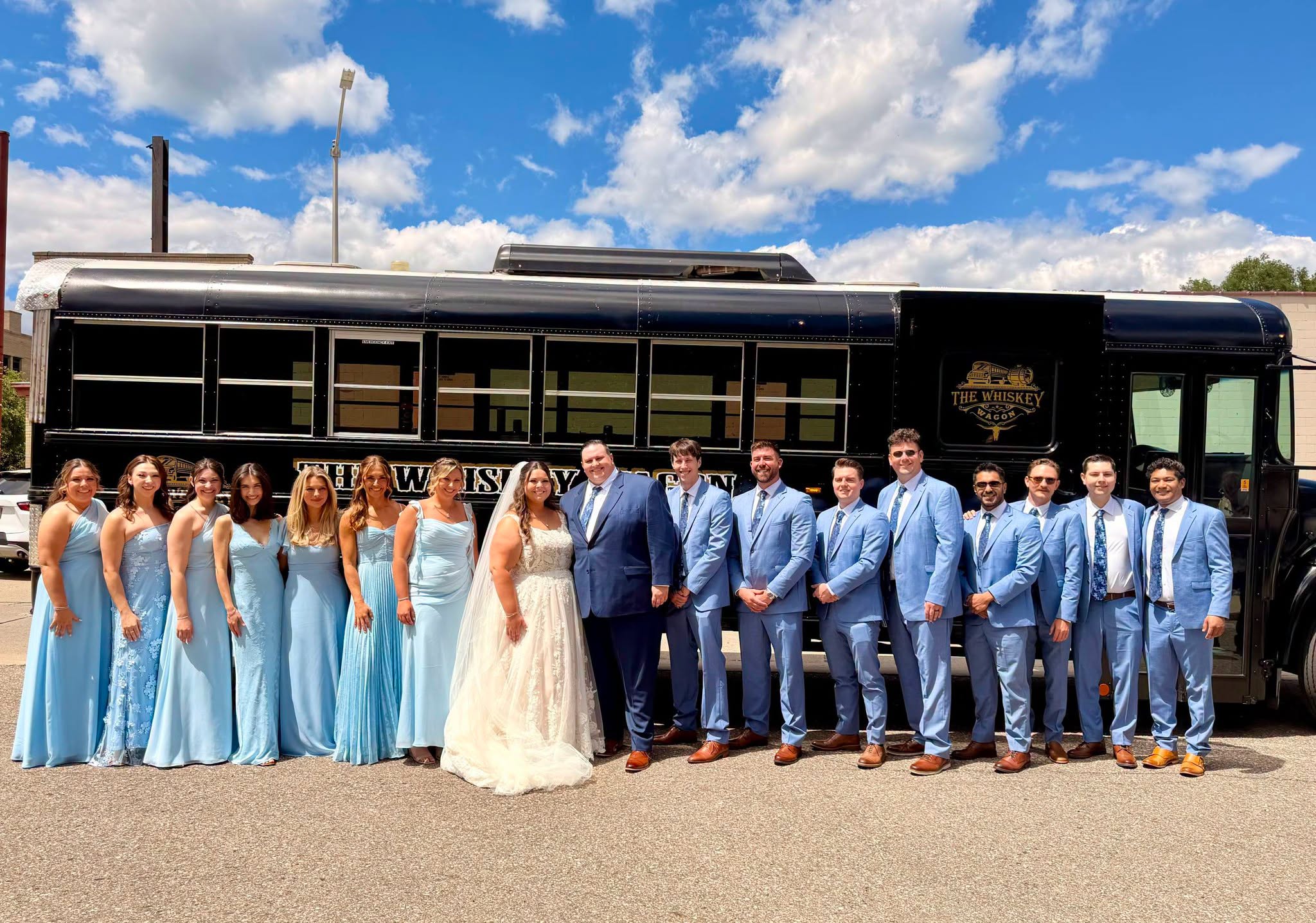 Group of people in formal attire standing in front of a black bus outdoors, with blue sky and clouds above.