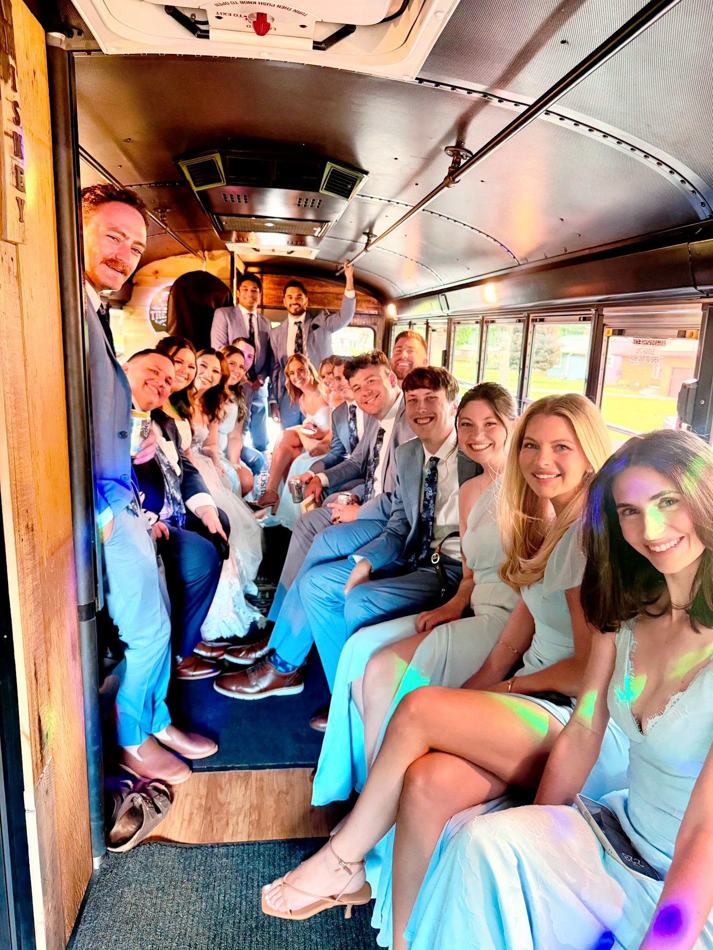Group of young people sitting inside a bus, smiling and posing for a photo, with sunlight coming through the windows.