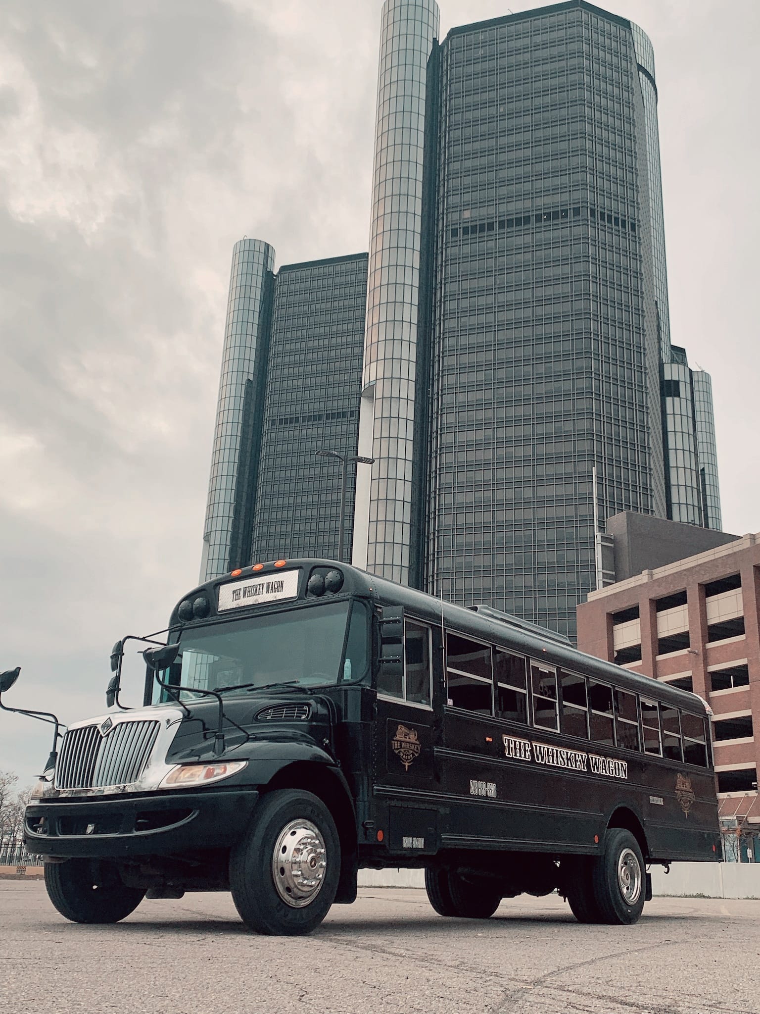Black school bus parked in front of tall modern glass skyscrapers, cloudy sky overhead.