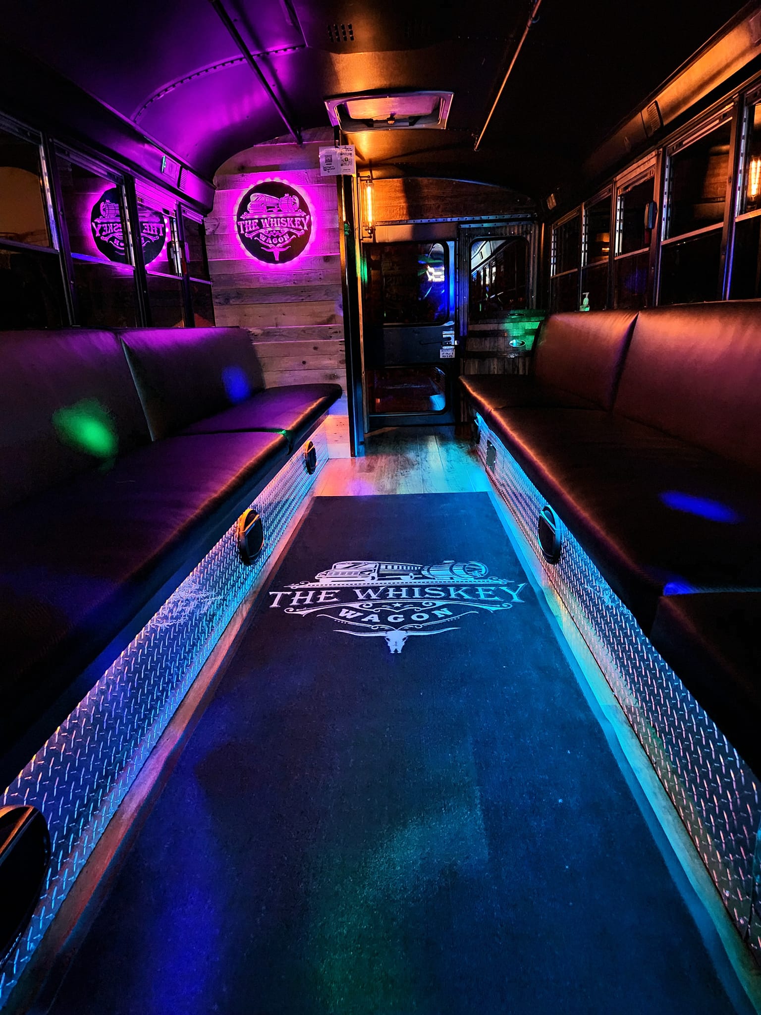 Interior of a bus or train with illuminated purple and blue lights, empty seats on both sides, and a logo on the floor.