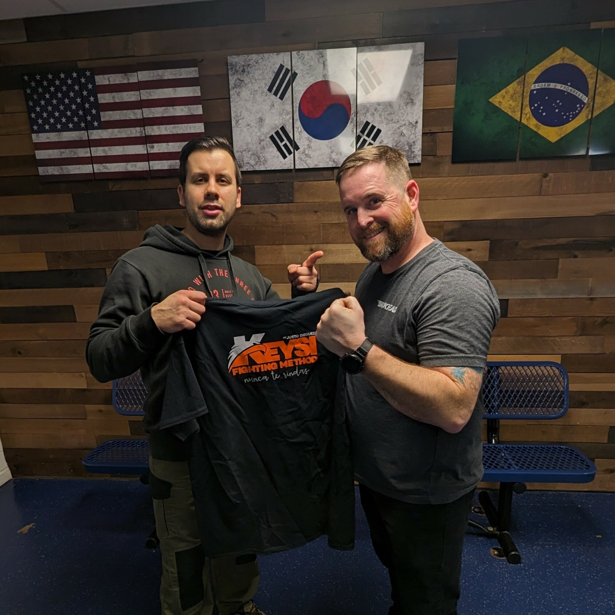 Two men standing indoors, holding a black T-shirt with orange text, smiling, with flags of the United States, South Korea, and Brazil in the background.