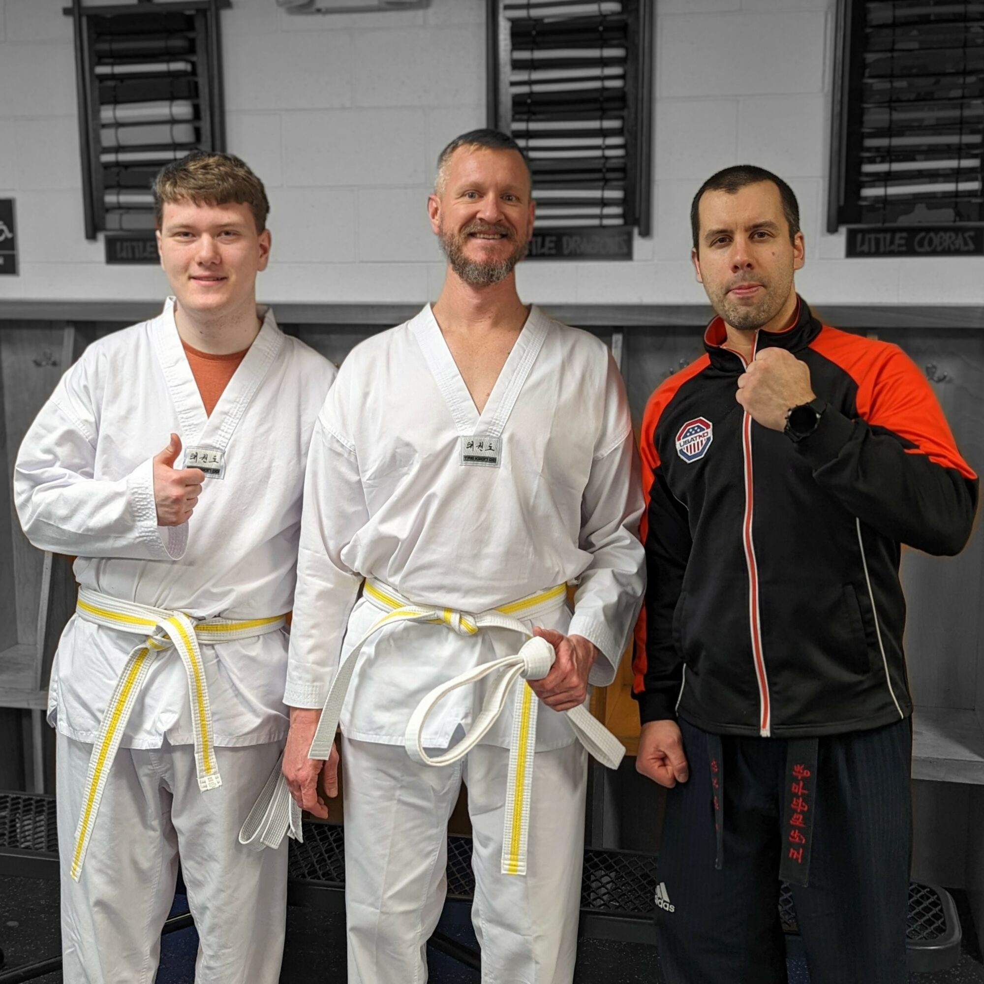Three men in martial arts uniforms and a jacket standing indoors, smiling, with black window blinds behind them.