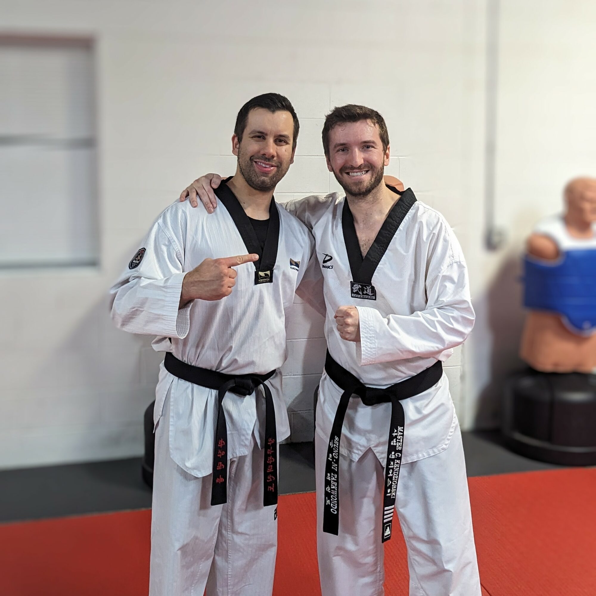 Two men in taekwondo uniforms smiling, with one pointing at the other, in a martial arts training area.