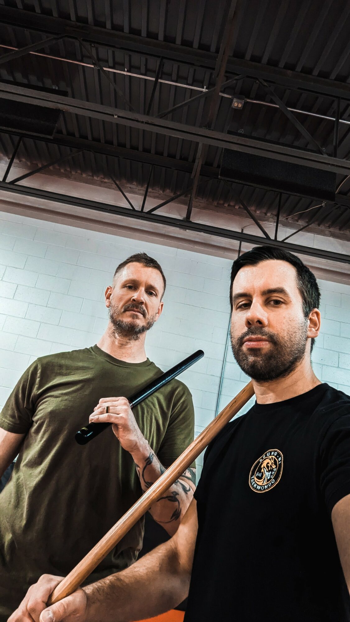 Two men holding pool cues in an indoor setting with a high ceiling and metal beams.