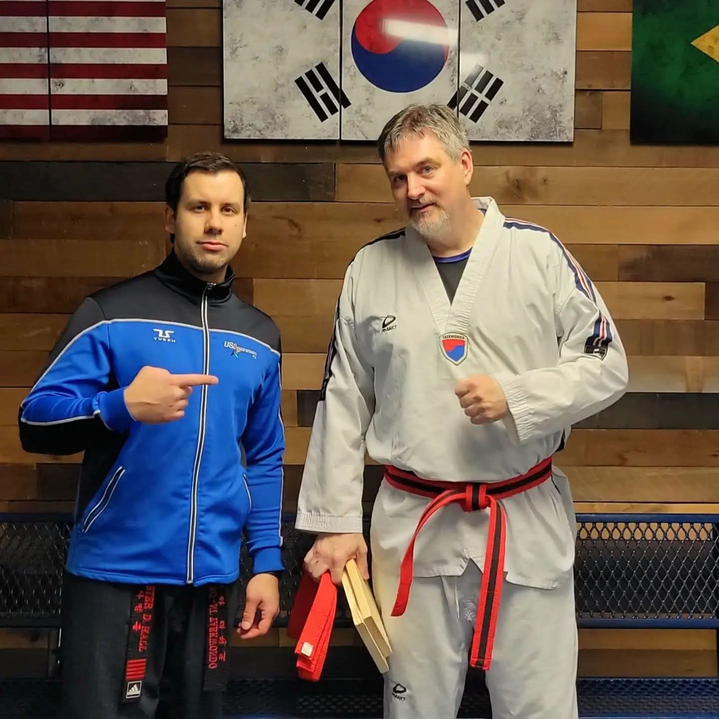 Two men standing indoors, one in a blue jacket and the other in a martial arts uniform with a red belt, holding a wooden board.