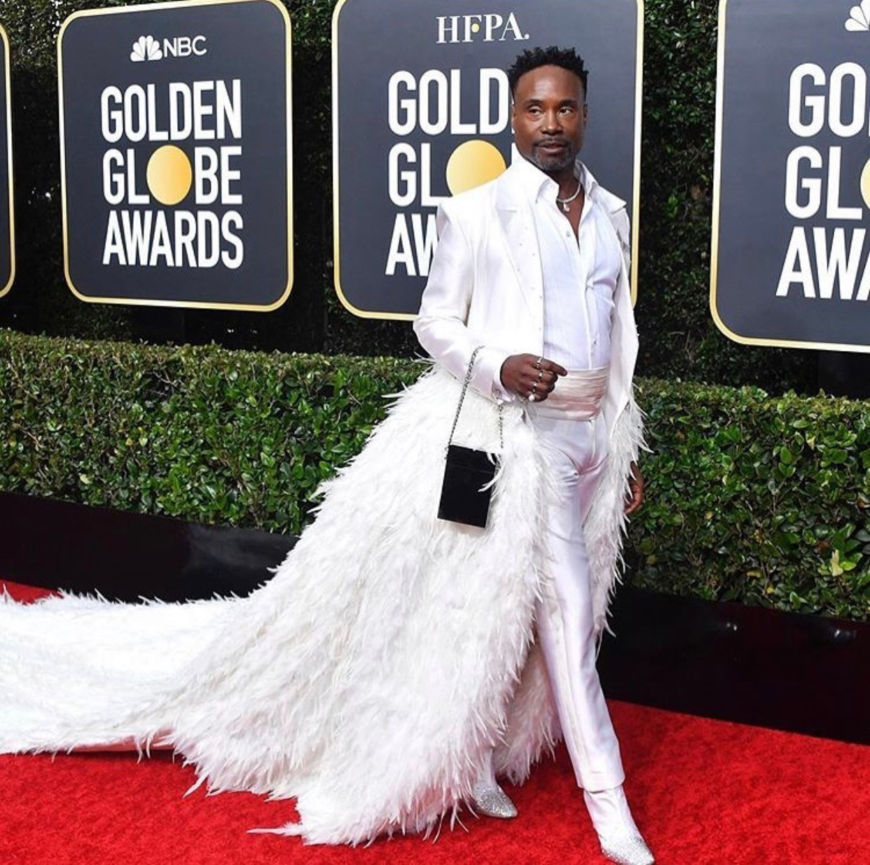 Person in white outfit with a long feathered train on red carpet at awards event, background with logos.