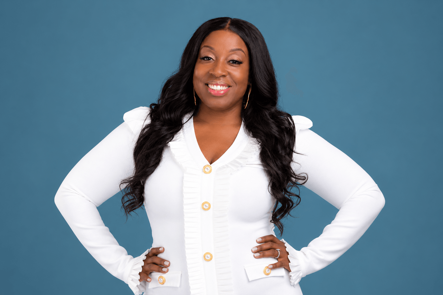 Woman with long dark hair smiling, wearing a white button-up shirt, hands on hips, against a blue background.