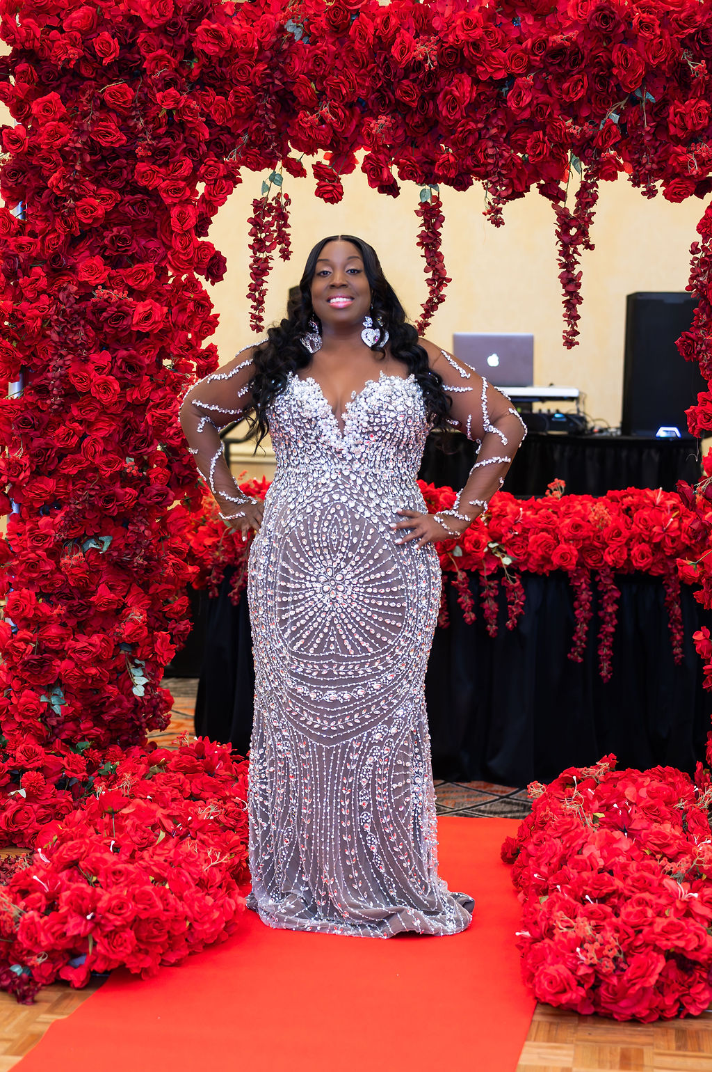 Woman in a silver, beaded gown standing on a red carpet surrounded by red flowers and floral arch.