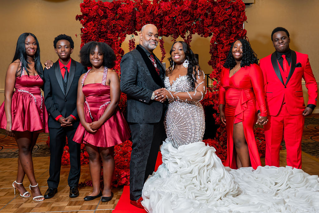 Group of seven people dressed in formal attire, standing in front of a red floral backdrop, smiling and holding hands.