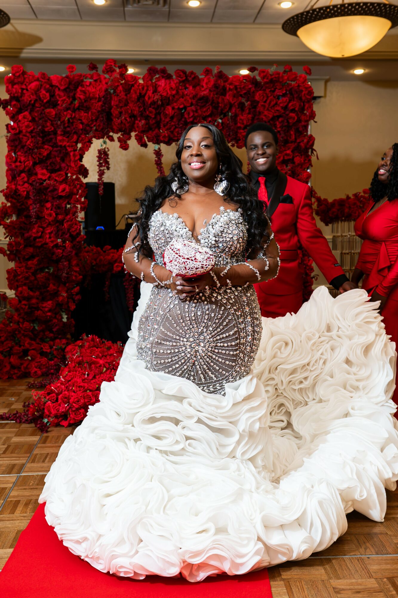 Woman in a wedding dress standing in front of a red floral backdrop, smiling, with a man in a red suit behind her, and a white ruffled train on the floor.