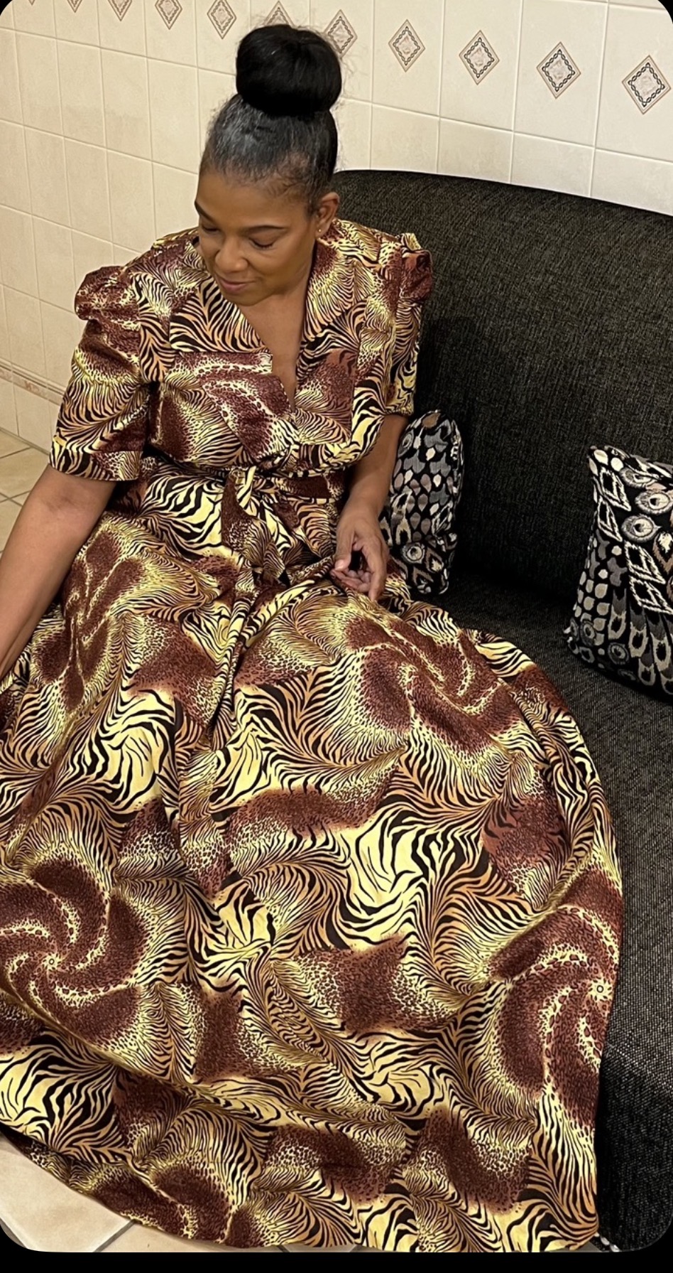 Woman in a patterned dress sitting on a black couch, looking down at her dress, with a black and white bag nearby.