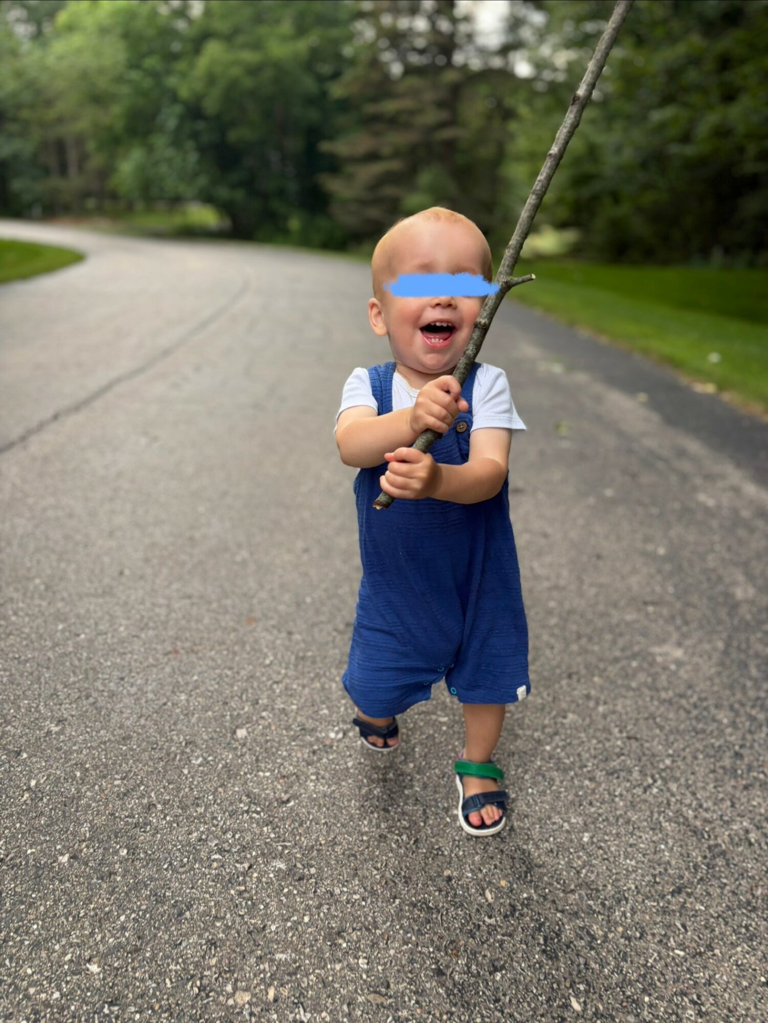 Child walking on a paved road holding a fishing rod, smiling, with trees in the background.