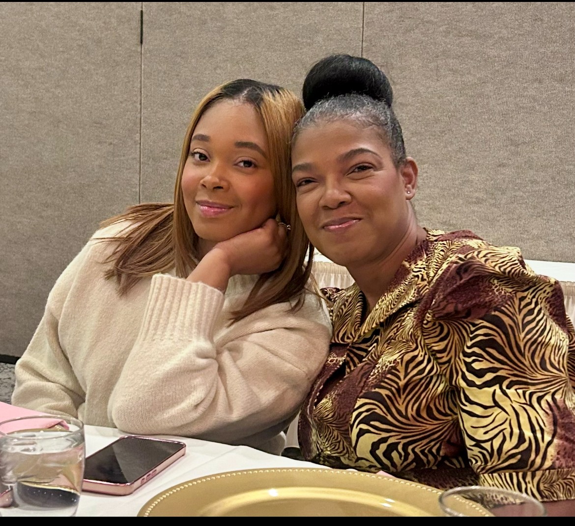 Two women sitting at a table, smiling, with a beige background. One woman has long hair, the other has her hair in a bun. They are close together, with one resting her head on her hand. On the table are a glass, a phone, and a plate.