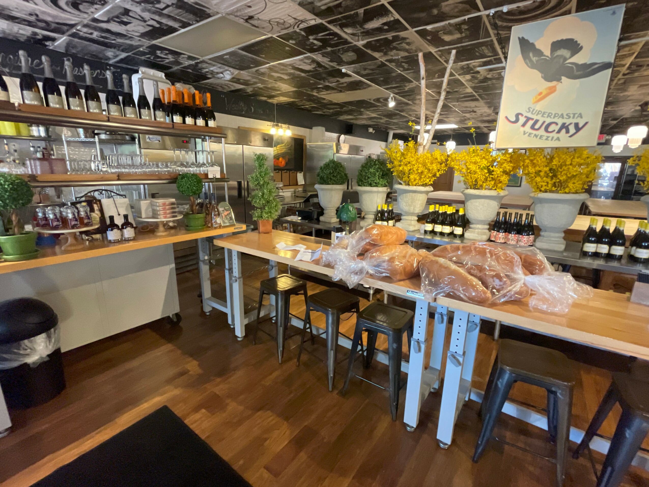 Interior of a cafe with tables, chairs, shelves with bottles, and yellow flowers, with a poster on the ceiling.