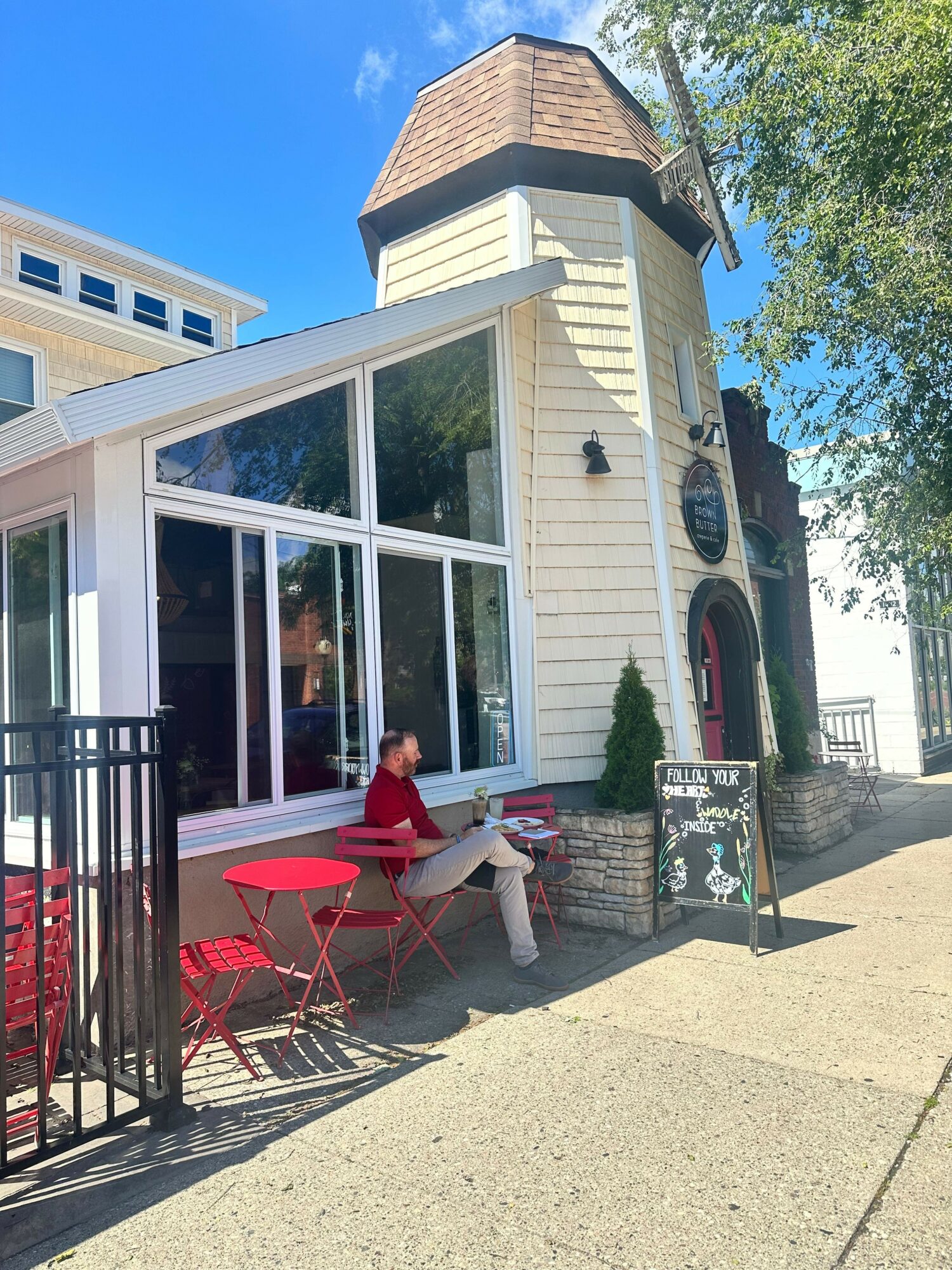 Person sitting at an outdoor table outside a small building with large windows, trees, and a sidewalk.