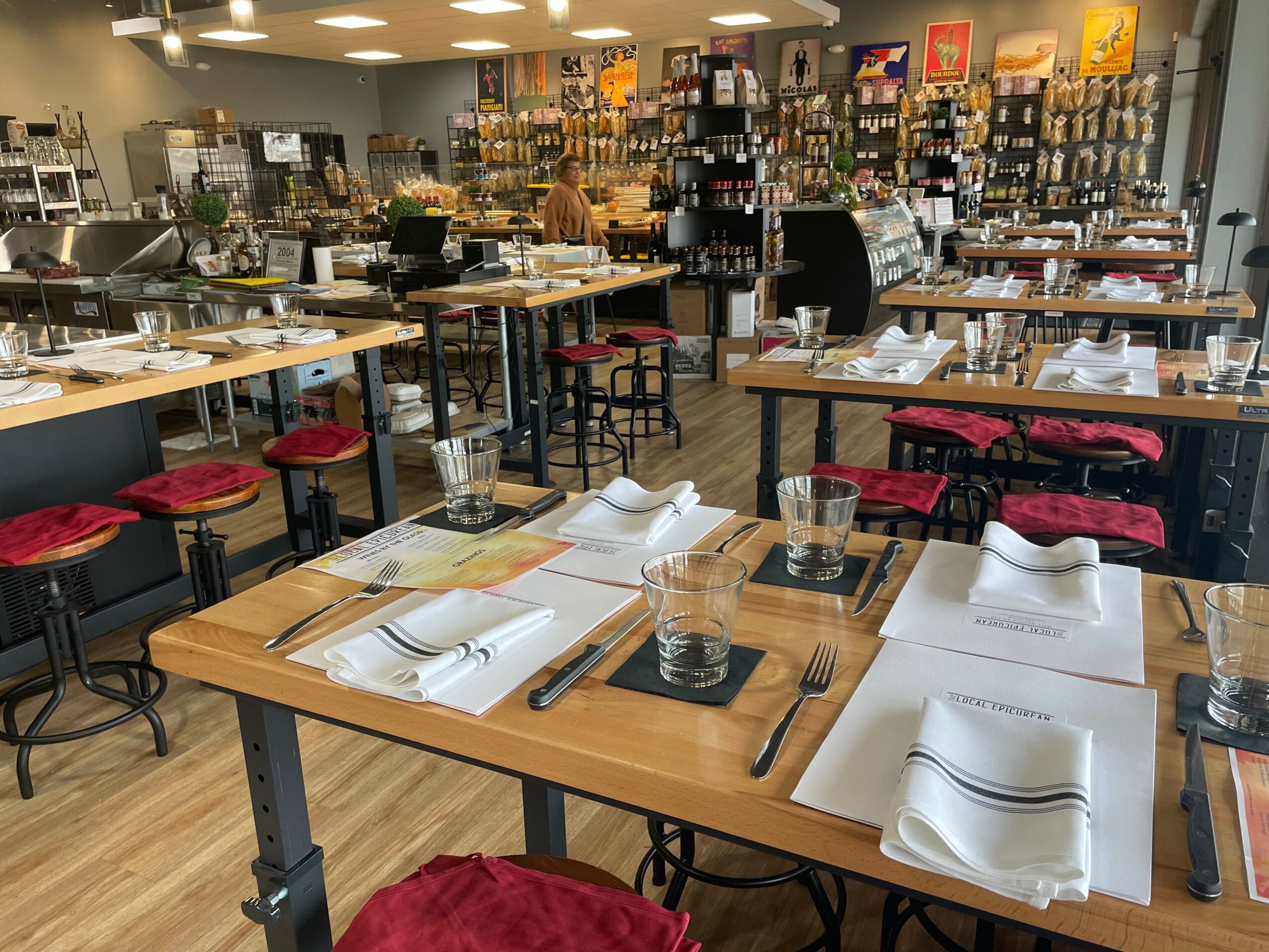Restaurant or cafe interior with tables set with napkins, glasses, and utensils, and shelves with products in the background.