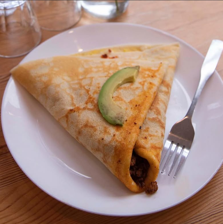 Folded crepe with avocado slice on white plate, fork on side, wooden table background.