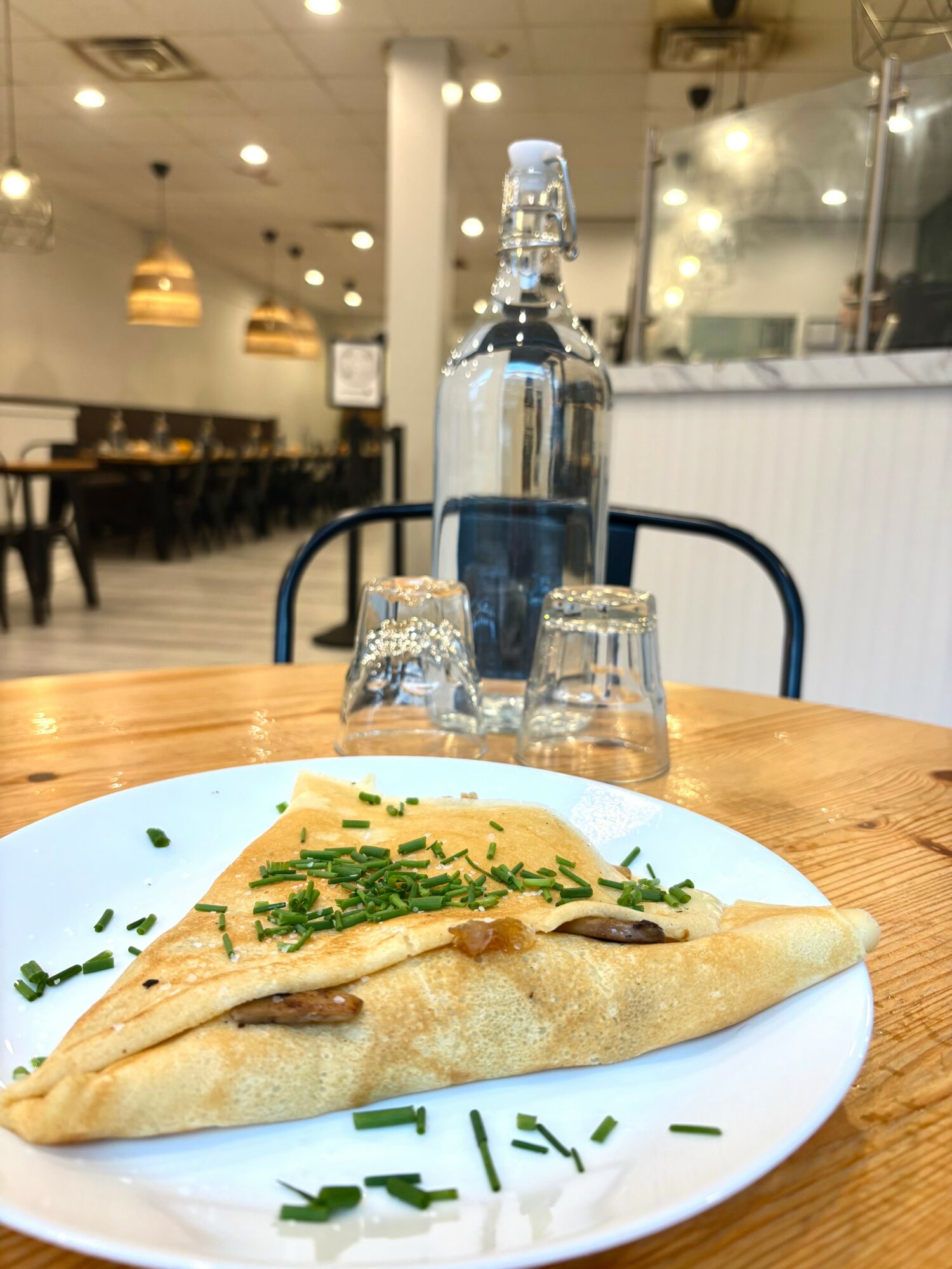 A folded crepe garnished with chopped chives on a white plate, with a glass water bottle and two glasses behind, in a cafe.