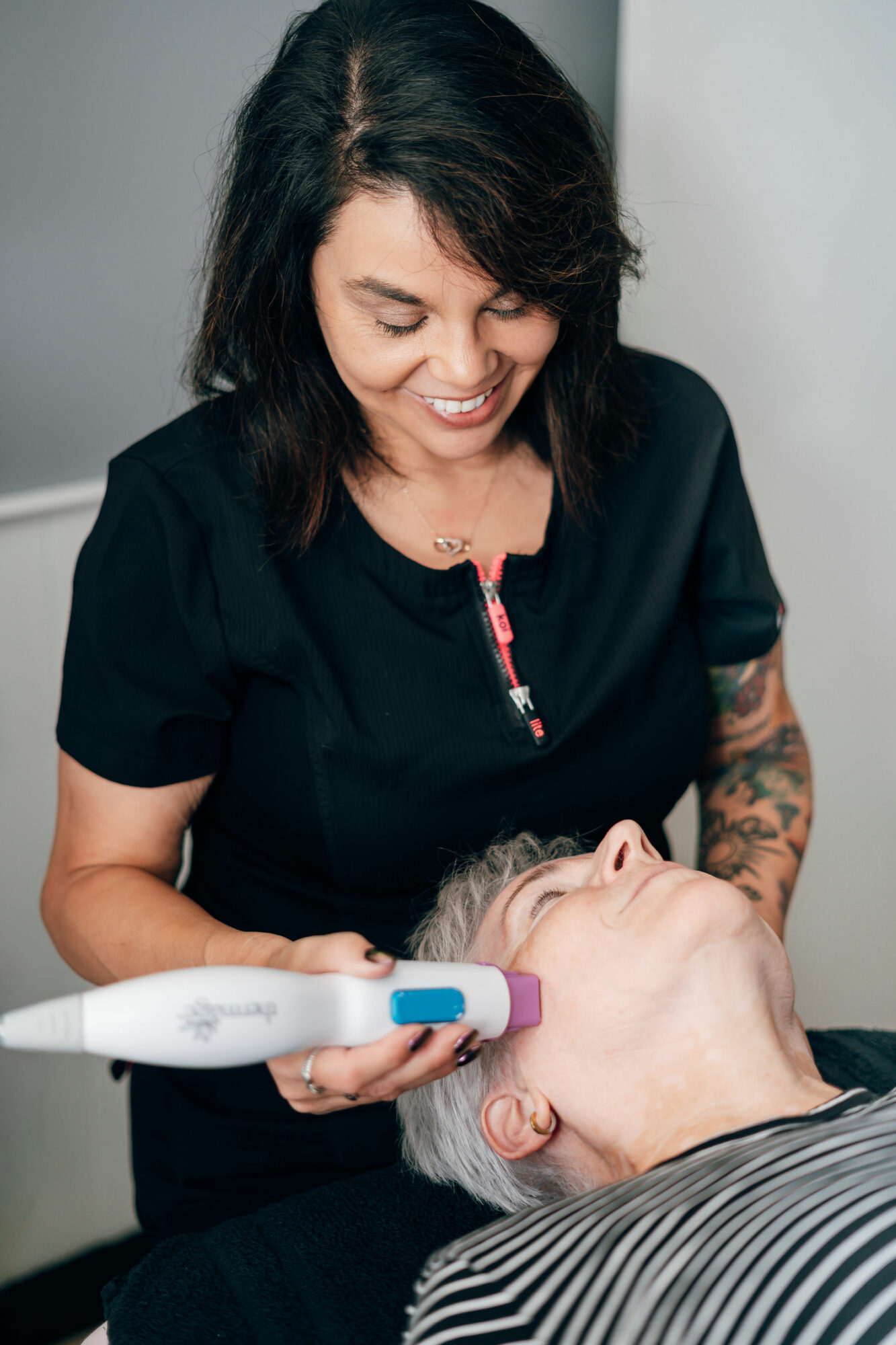 A woman with dark hair smiling while holding a device near an older person's face in a clinical setting.