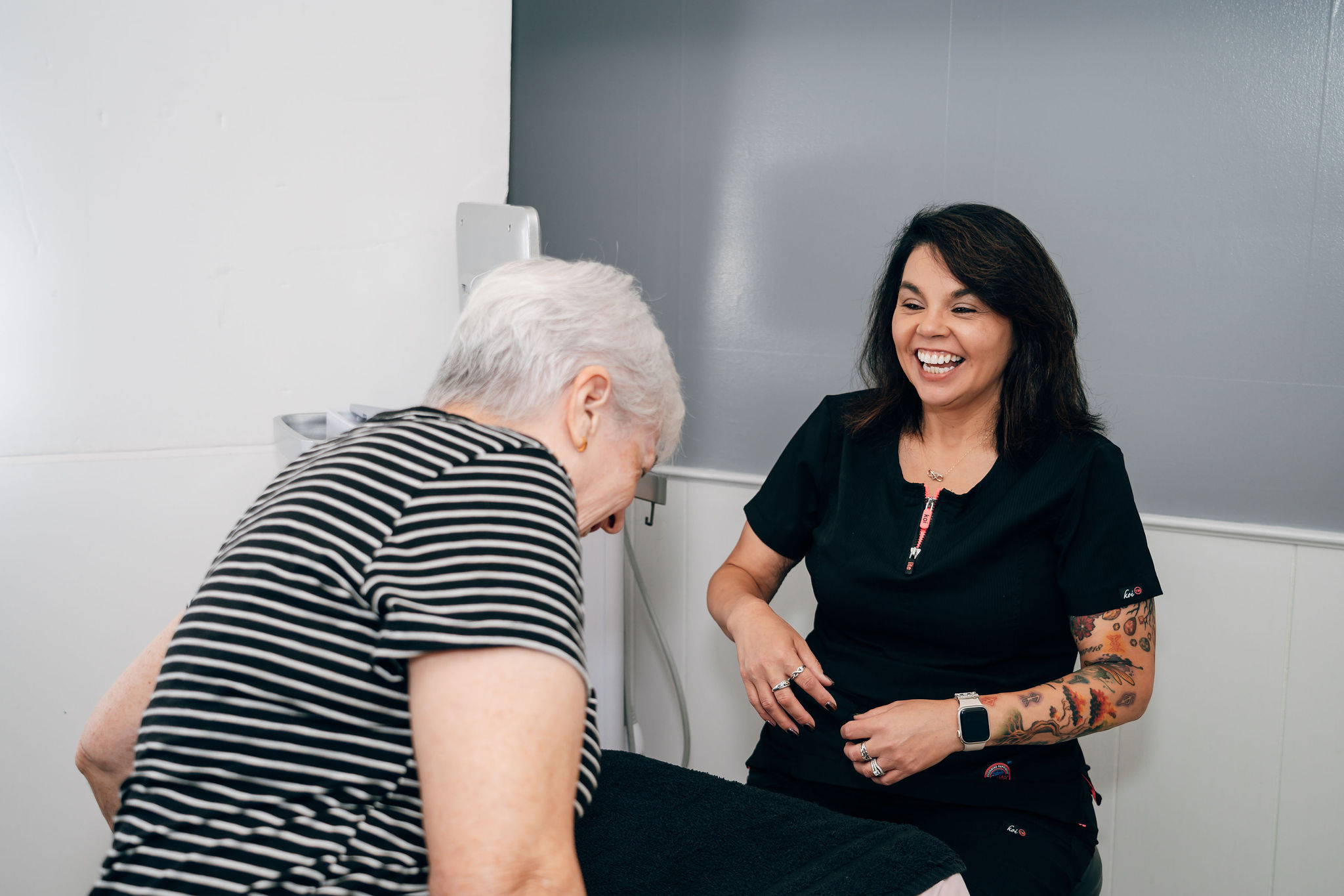 Two women laughing and talking in a modern indoor setting, one with dark hair and tattoos, the other with gray hair.