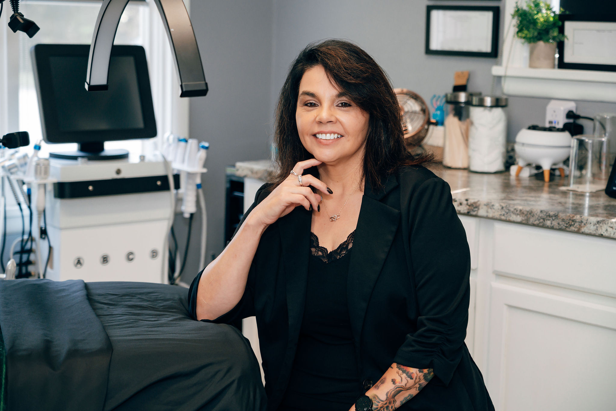 Smiling woman with dark hair and tattoos in a medical or dental office, sitting near equipment and supplies.