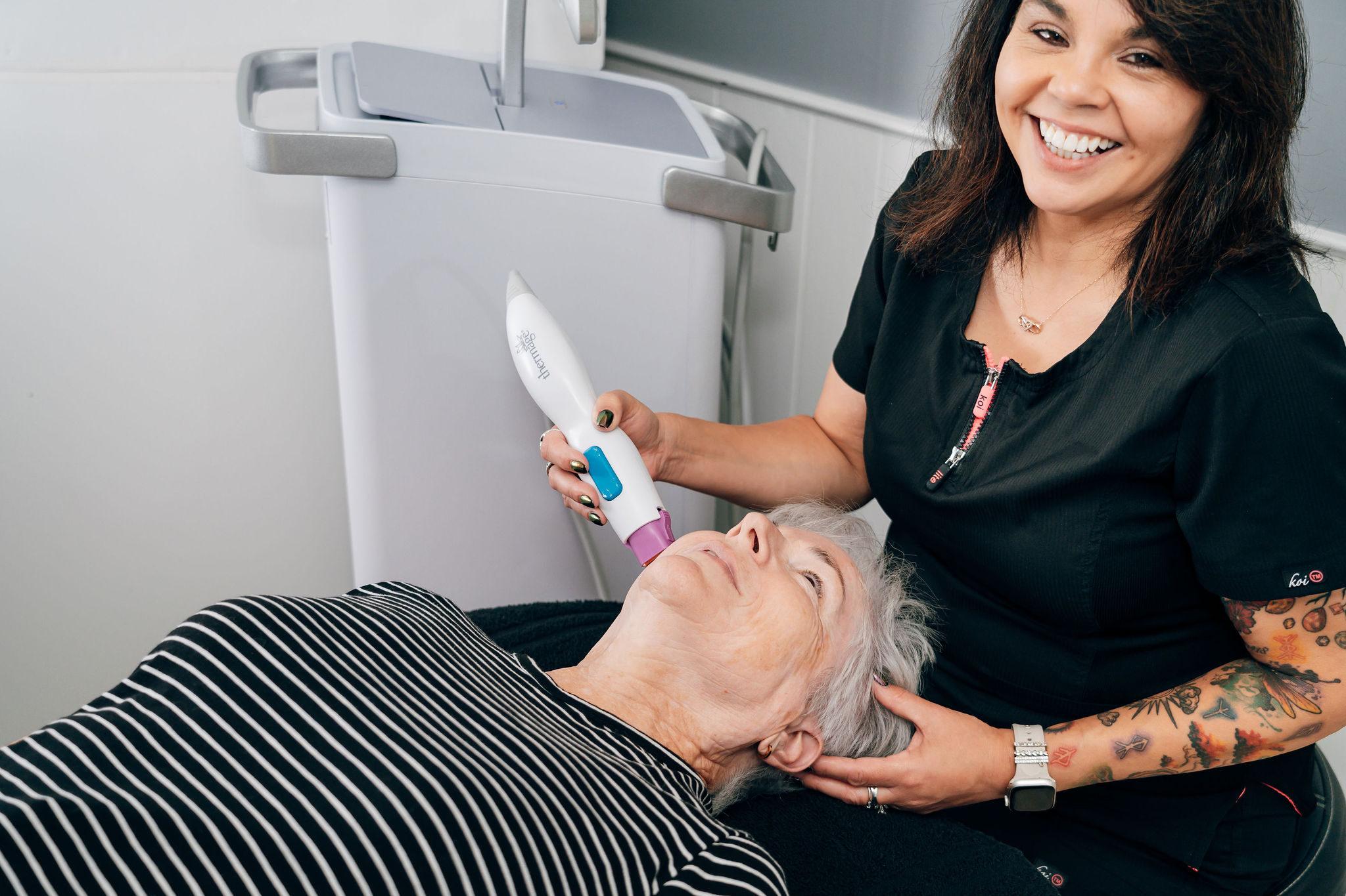 A woman smiling while holding an elderly person's head during a medical or dental procedure, with a large white machine in the background.