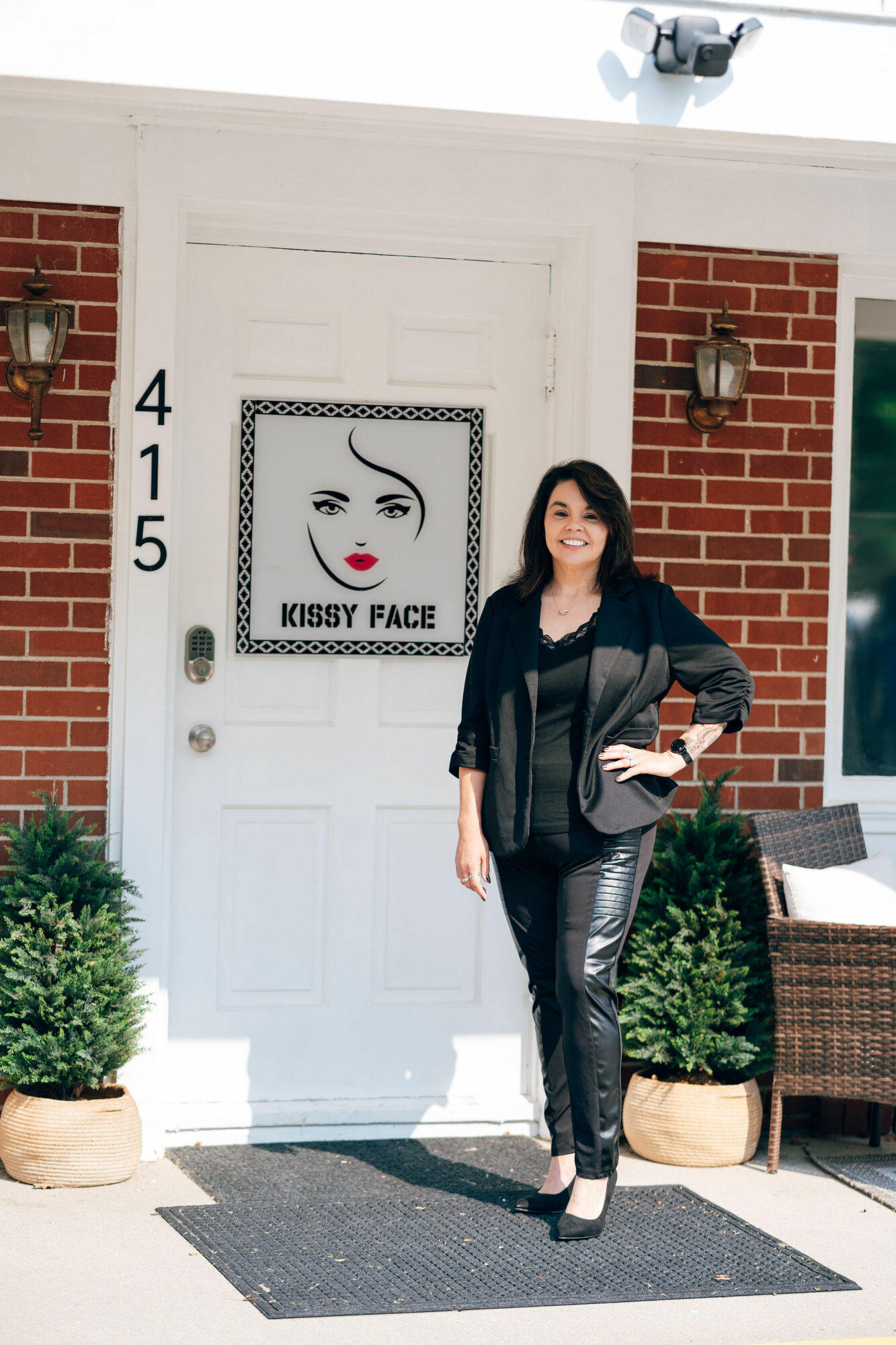 Woman standing outside a door with a sign reading 'KISSY FACE' and a drawing of a face, smiling, with brick and white wall background.