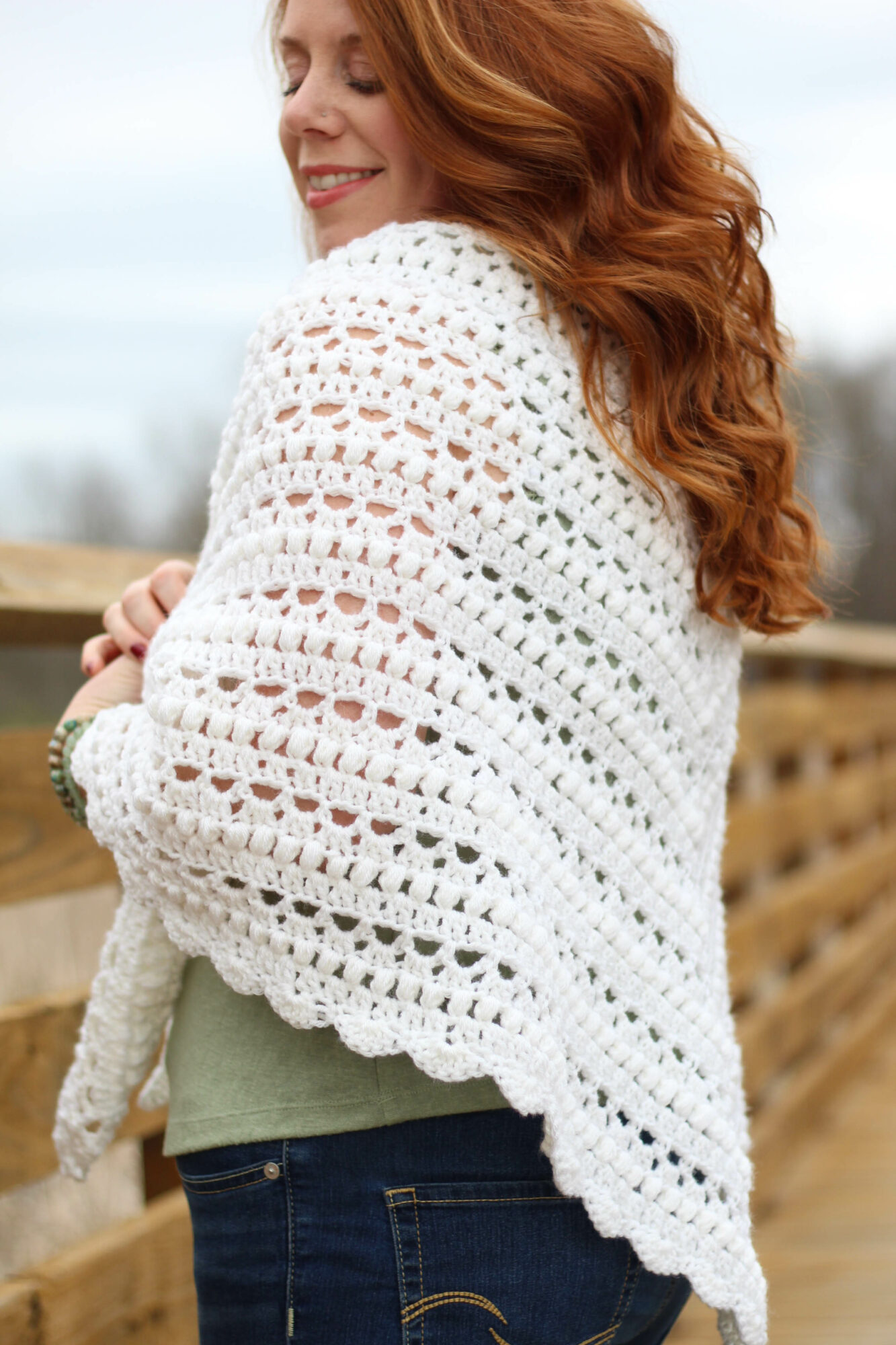 Woman with long, curly red hair wearing a white crochet shawl outdoors, smiling, with a wooden fence in background.