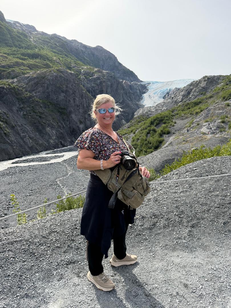 Woman with sunglasses holding a camera and backpack, standing on rocky terrain near a waterfall and mountain landscape.