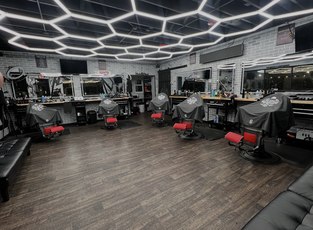 Barber shop with chairs covered in black capes, mirrors, and workstations, illuminated by geometric ceiling lights, with a wooden floor.