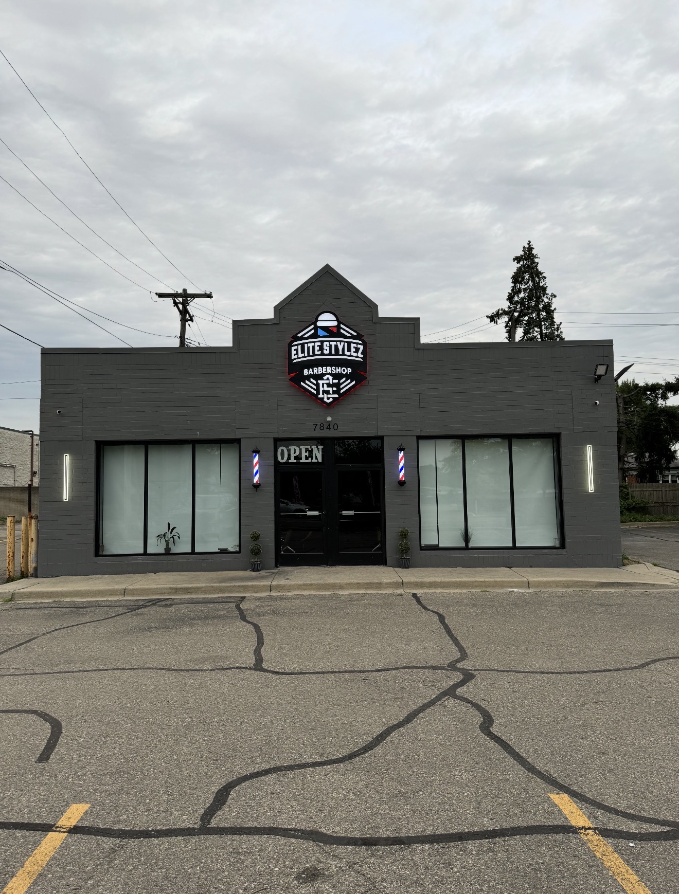 Gray building with a sign reading 'Elite Stylez' and an 'Open' sign, two large windows, and a parking lot in front.