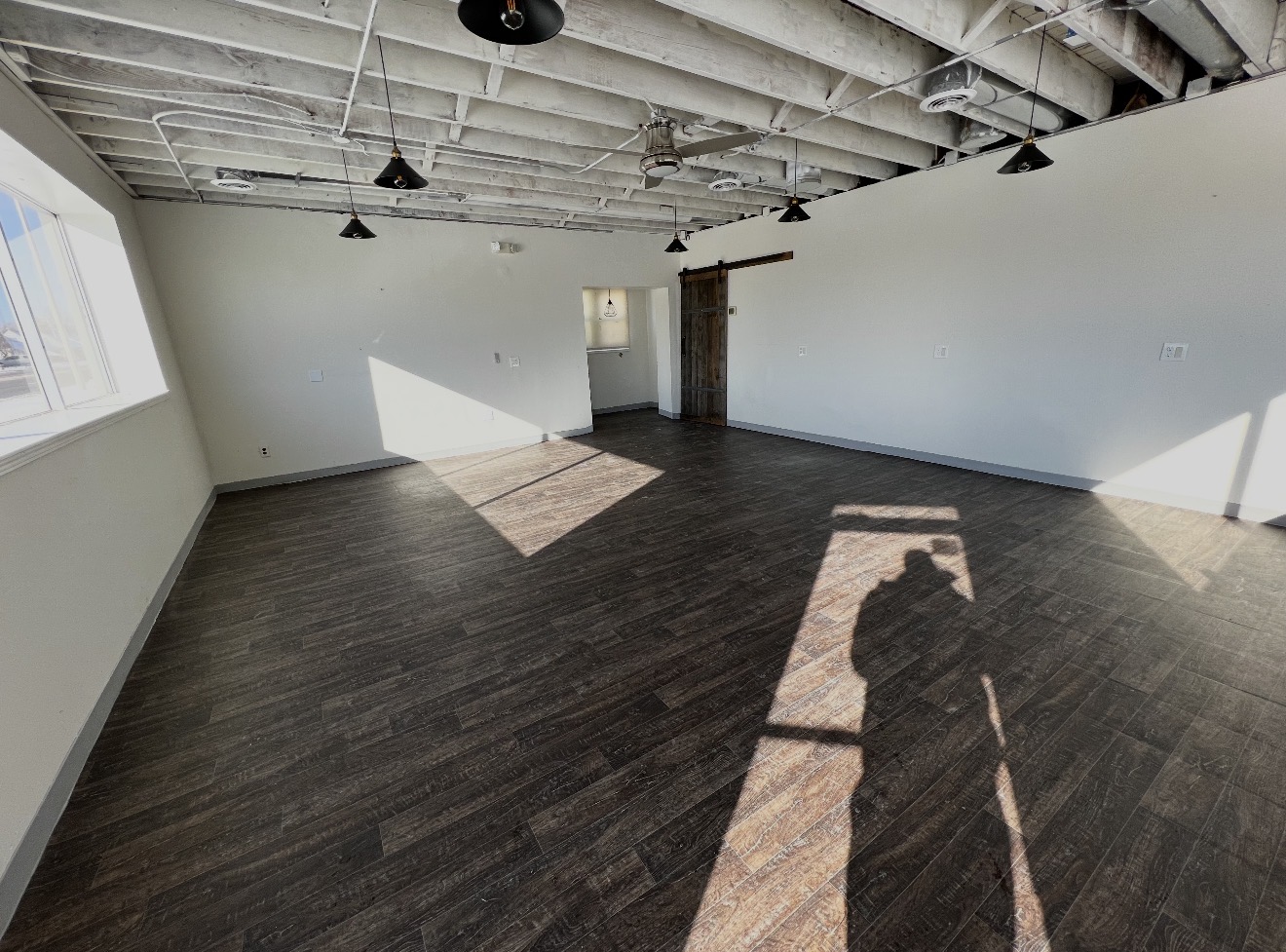 Empty room with white walls, dark wood floor, large window, and exposed ceiling beams, sunlight casting shadows inside.